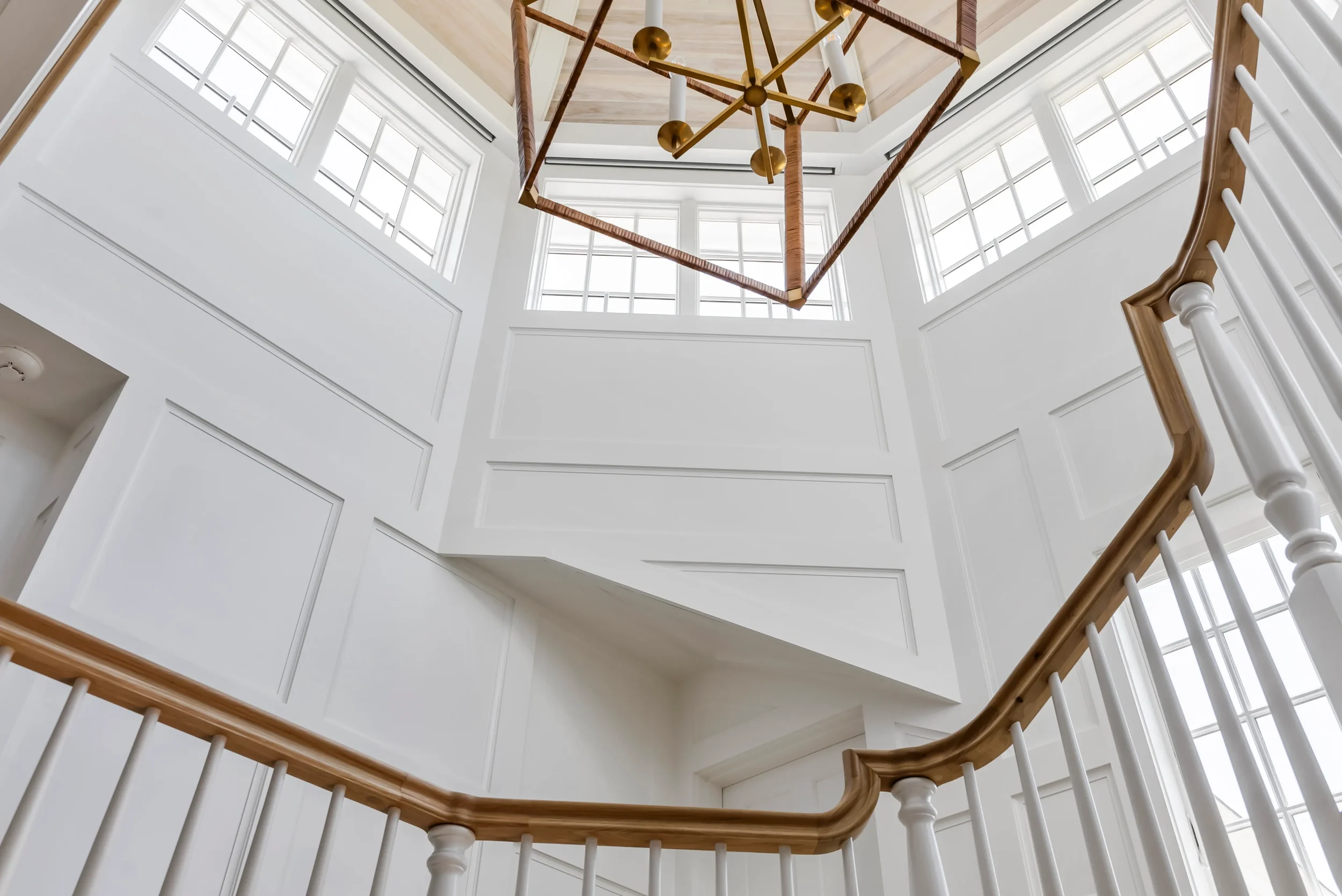 Interior view of a bright staircase with white walls, wooden handrails, and large windows at the top, featuring a modern geometric chandelier hanging from the ceiling.