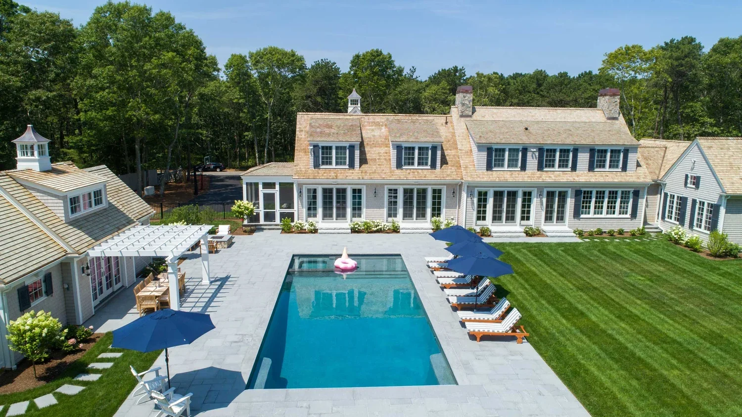A large backyard with a swimming pool, surrounded by a patio with lounge chairs and umbrellas, a well-maintained lawn, and a white house with multiple windows and a porch.