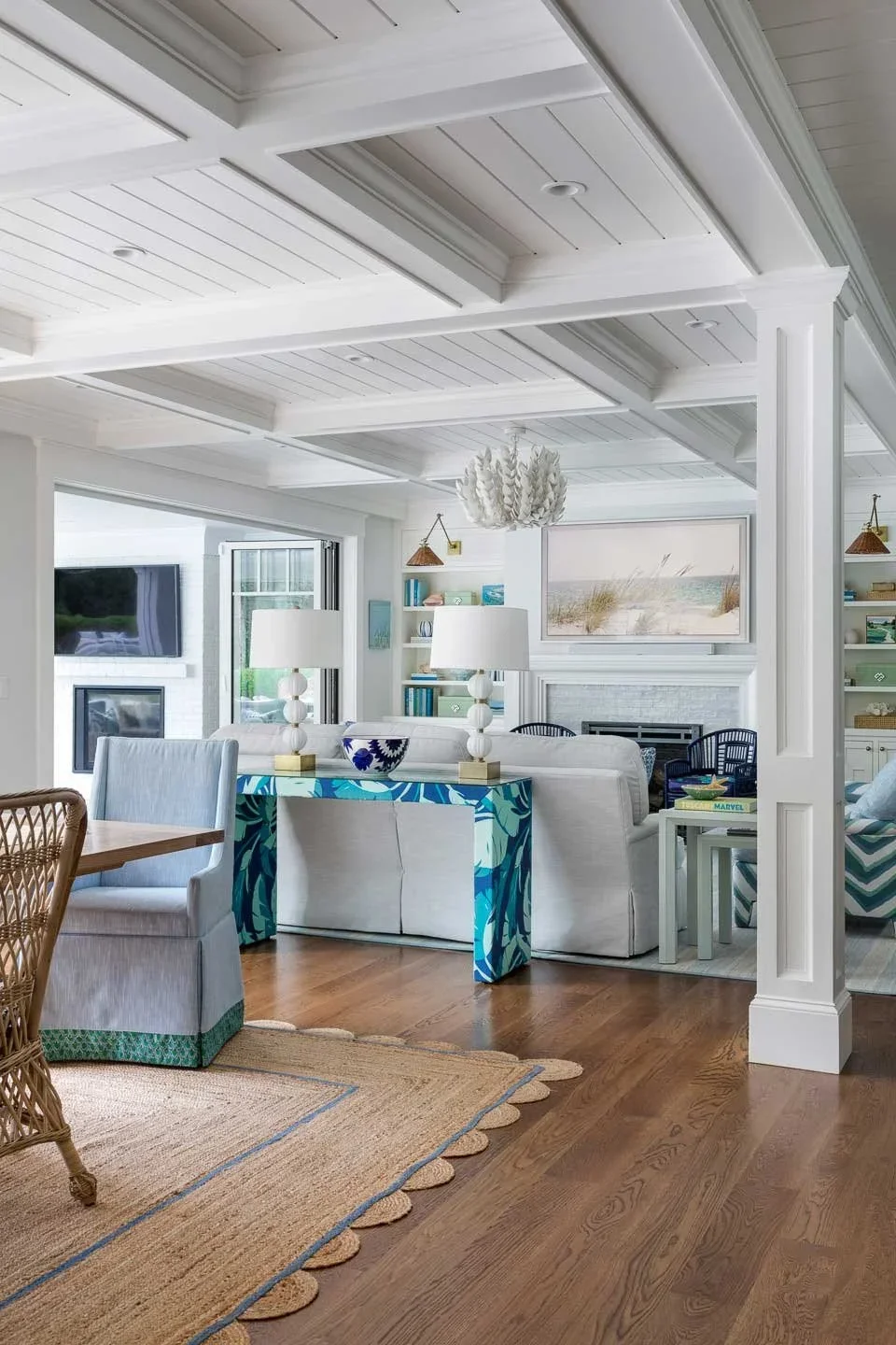 This image showcases a coastal-style living room that masterfully blends relaxed elegance with modern sensibility. The space is anchored by white coffered ceilings with recessed lighting, crisp white walls, and warm hardwood flooring—all contributing