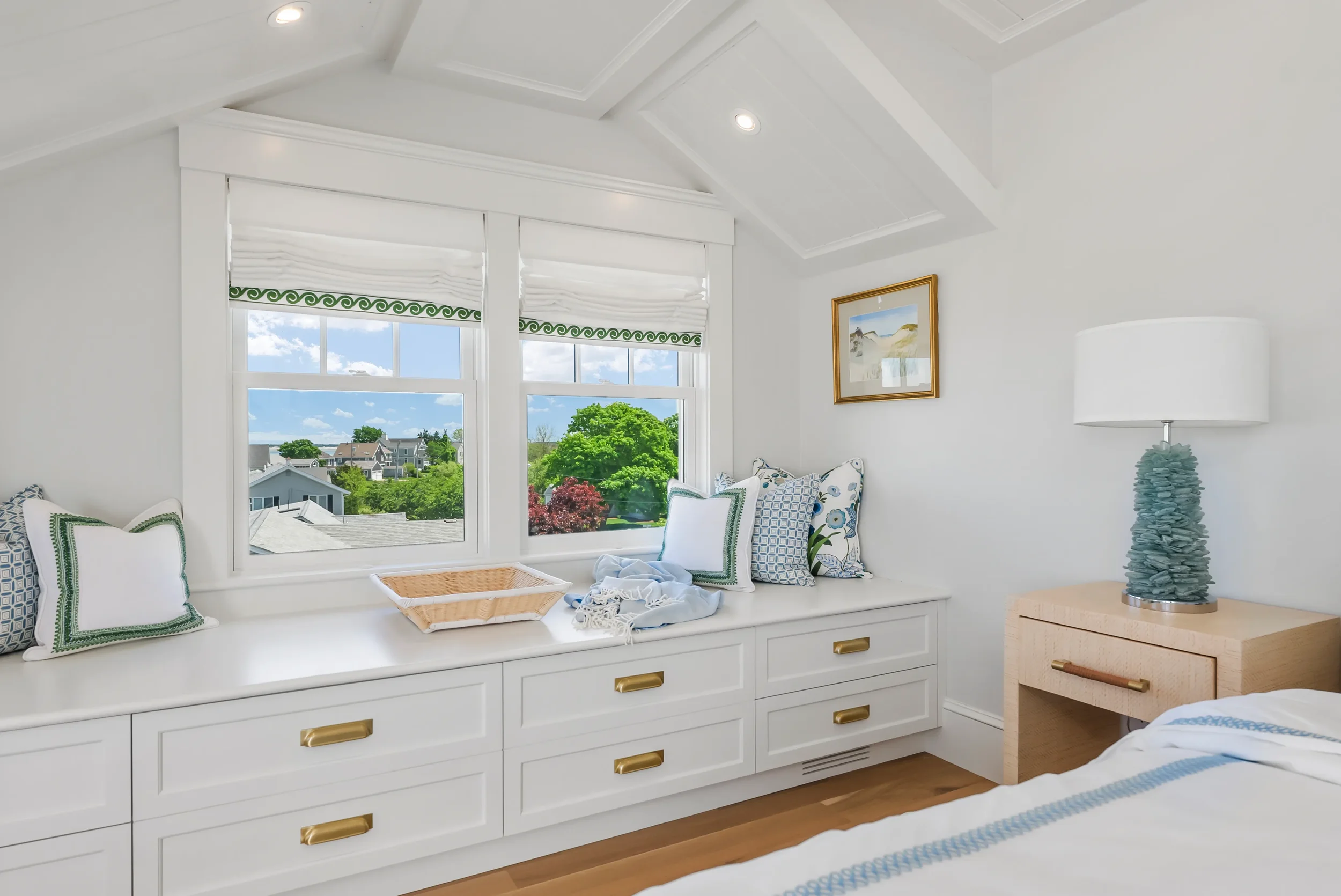 Bright bedroom with a large window seat, decorated with blue and white pillows, overlooking a green landscape and blue sky.