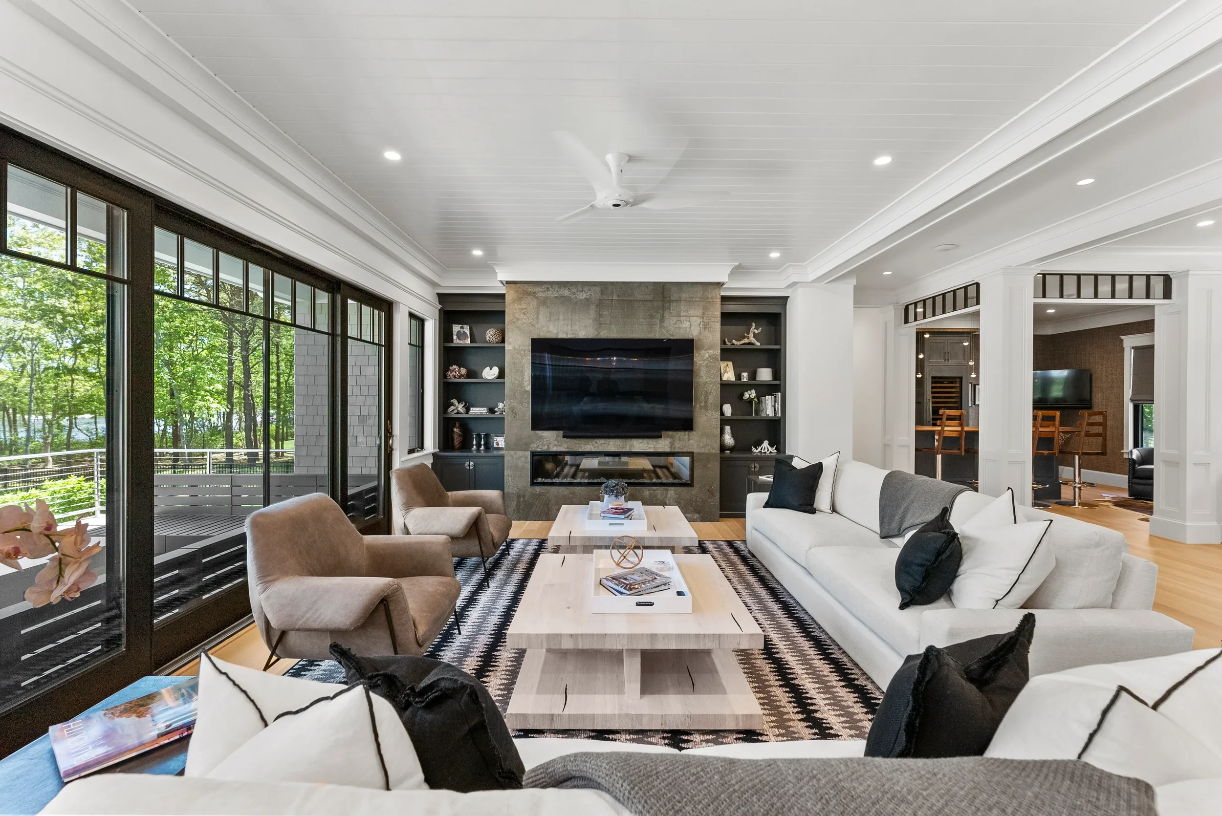 Modern living room with white sofa, brown chairs, large flat-screen TV, black built-in shelves, glass sliding doors showing trees outside, and a patterned rug.