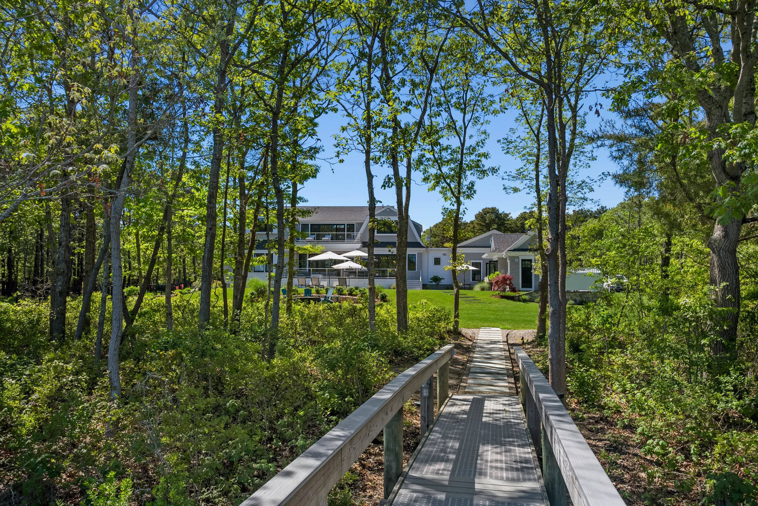 A wooden pathway leads through a wooded area to a spacious backyard with a house, patio, umbrellas, and outdoor furniture, under a clear blue sky.