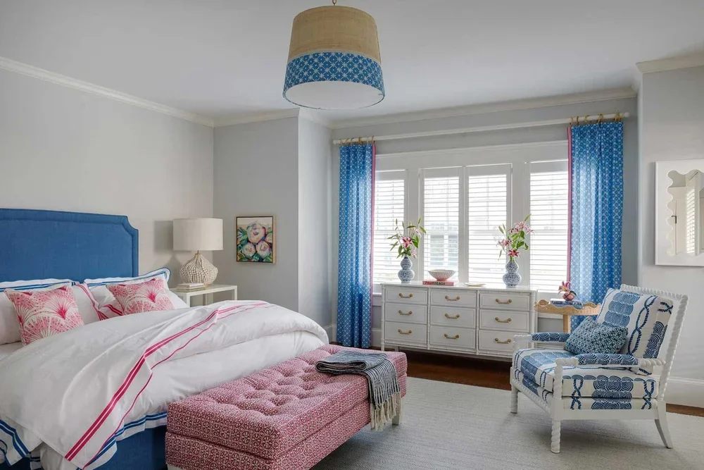 This image showcases a beautifully styled bedroom that blends elegance with comfort through a cohesive palette of blue, pink, and white. The bed features a blue upholstered headboard and crisp white bedding accented with pink details and patterned pi