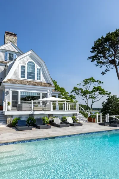 A house with a swimming pool in the backyard, with lounge chairs and trees around it, under a clear blue sky.