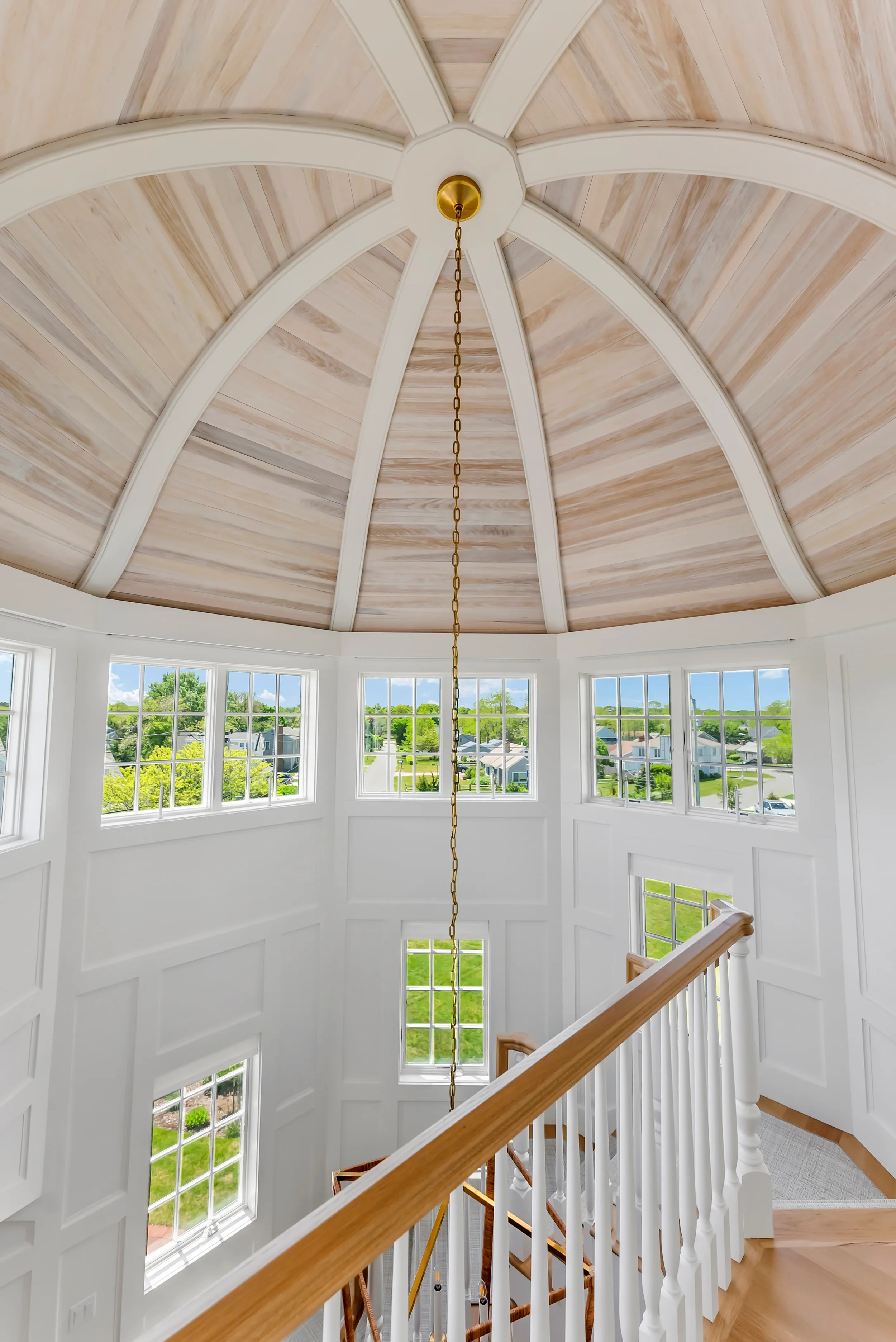 Interior view of a house's stairwell with large windows, white walls, a wooden handrail, and a vaulted ceiling with wood panels and white beams.