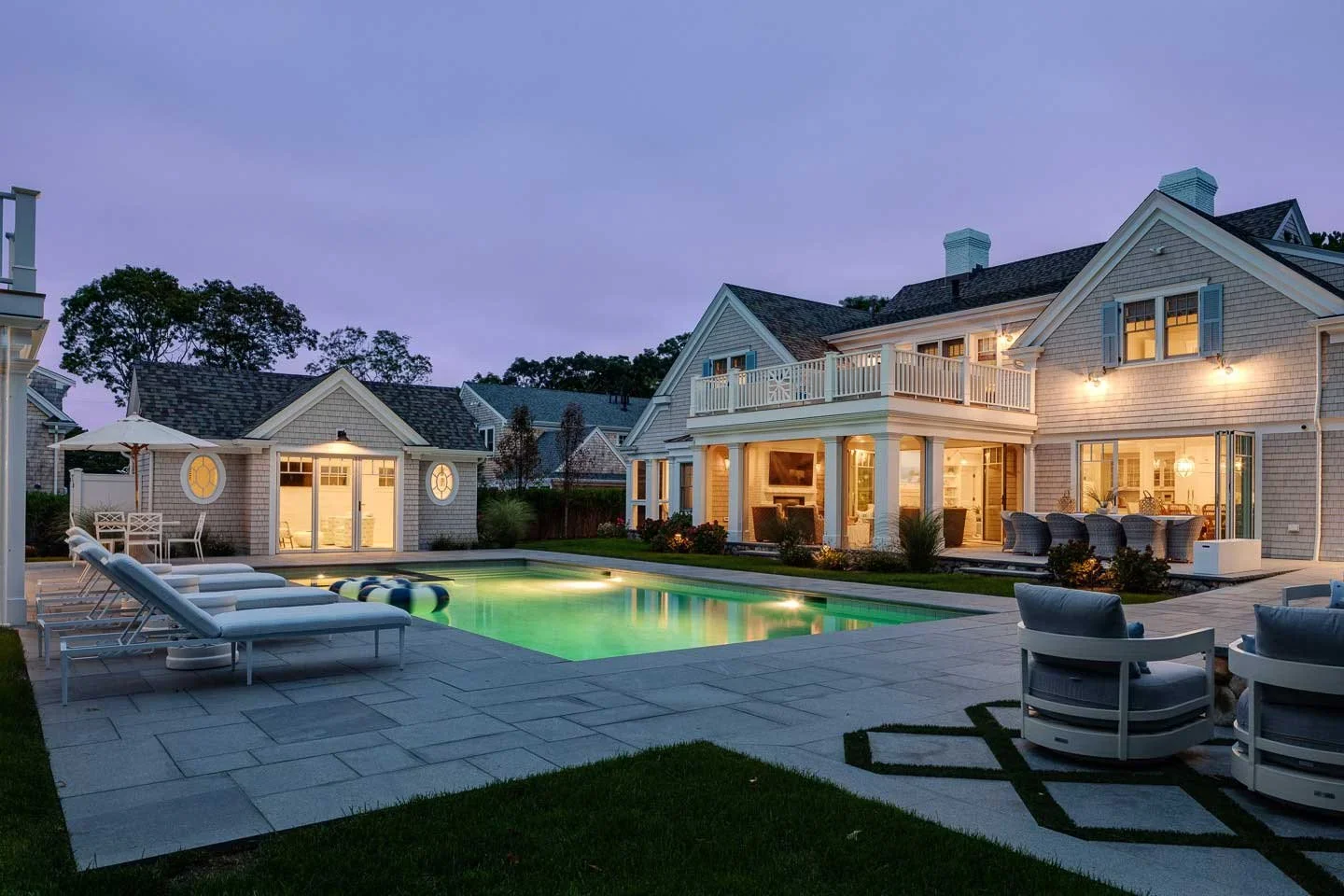 This image showcases a luxurious residential backyard at twilight designed for relaxation and upscale outdoor living. At the center is a large swimming pool, surrounded by a meticulously maintained stone patio and lush green lawn. Four white lounge