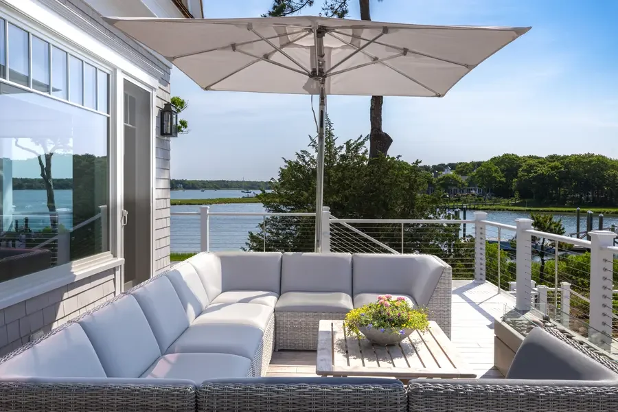 Outdoor balcony with white wicker sectional sofa and a coffee table with a flower arrangement, large outdoor umbrella, overlooking a body of water with trees and houses in the distance.