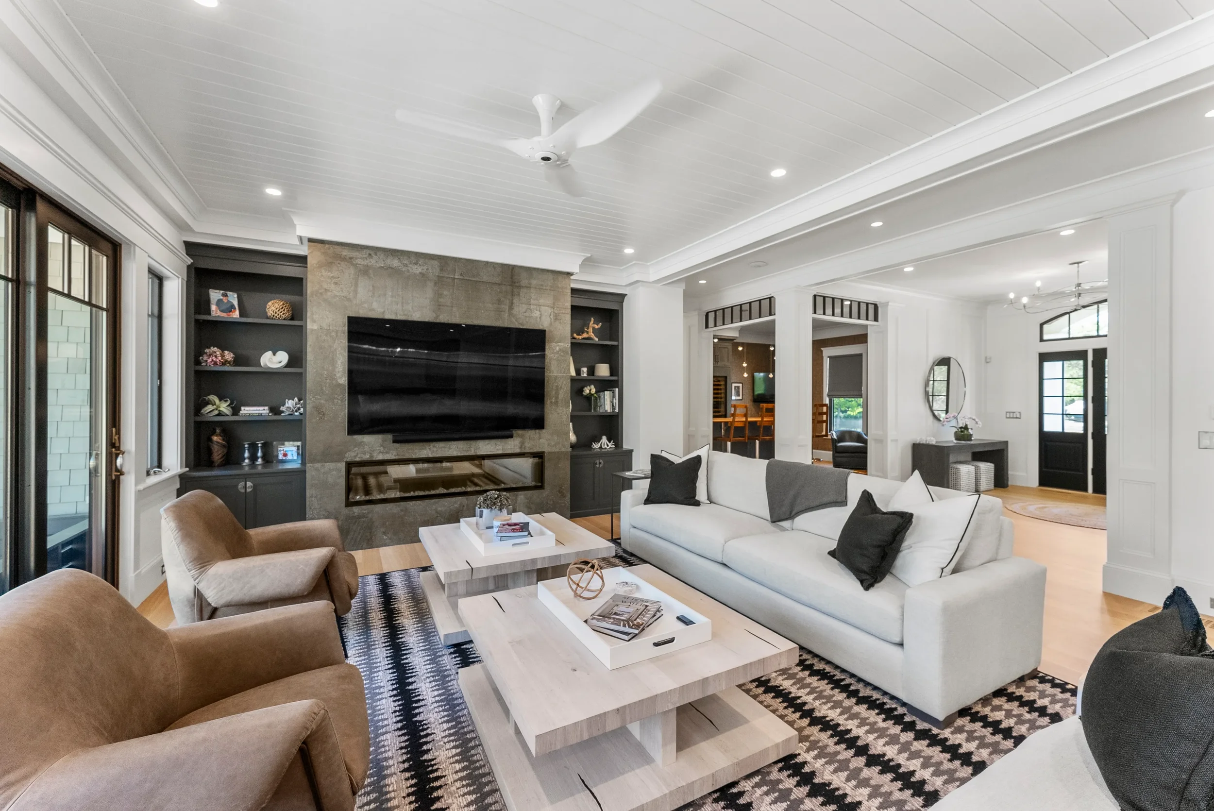 Modern living room with white walls, a white ceiling with recessed lighting, and wooden flooring. Features a large white sofa with black and gray pillows, a light wood coffee table, beige chairs, a black wall-mounted TV, built-in bookshelves, and a f