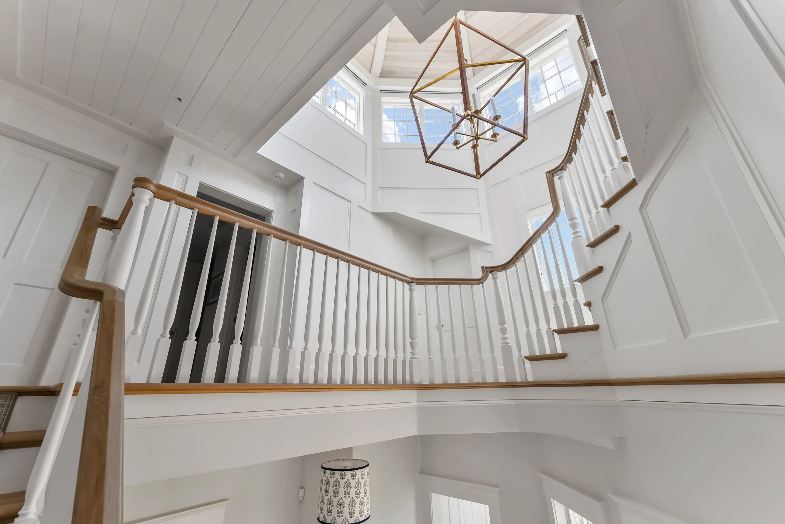 Interior view of a bright, spacious staircase with white walls, wooden handrails, and a geometric chandelier hanging from the ceiling, with large windows letting in natural light.