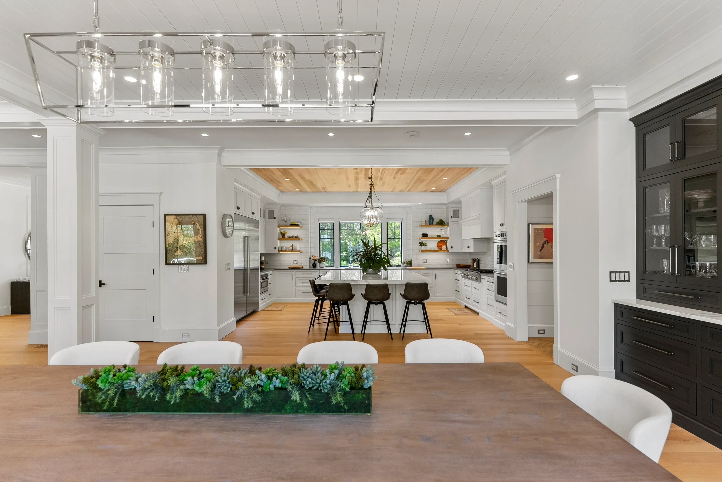 Spacious, modern kitchen with white cabinets, a large island with four chairs, open shelving, and a dining area with a wooden table and white chairs, decorated with a greenery centerpiece.