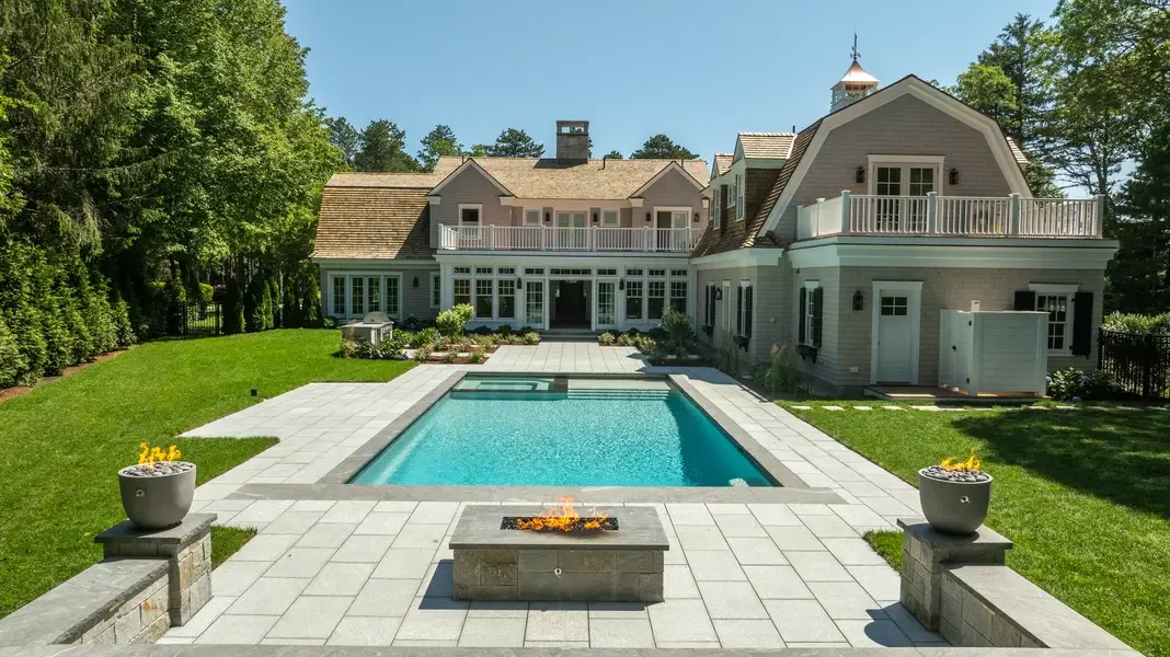 This image presents a magnificent luxury estate showcasing classic New England architecture arranged around a stunning pool courtyard. The expansive residence features weathered cedar shingle siding in soft gray tones complemented by crisp white trim