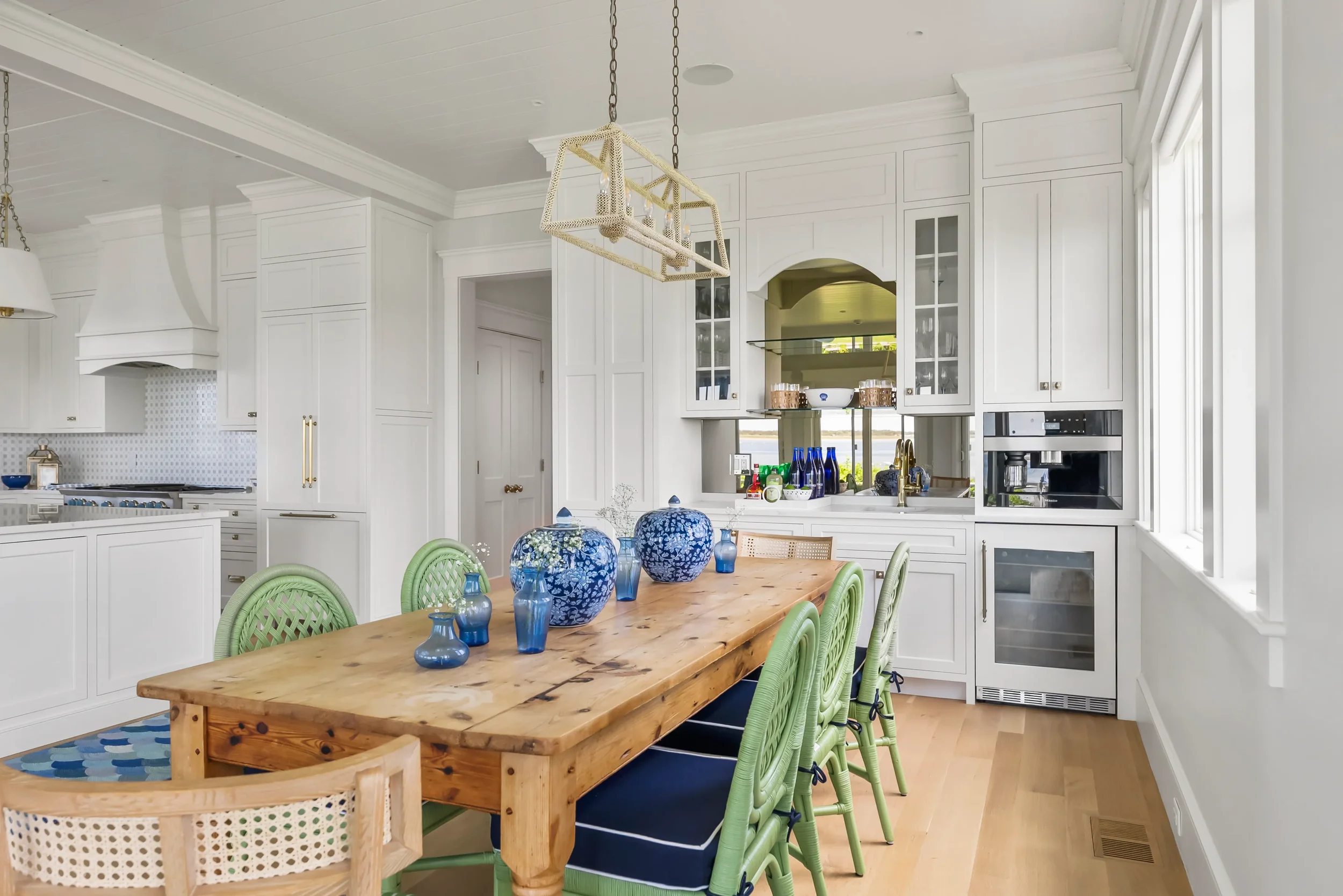 Bright white kitchen with a wooden dining table, green chairs, and blue vases and bowls as decor. Large windows let in natural light.