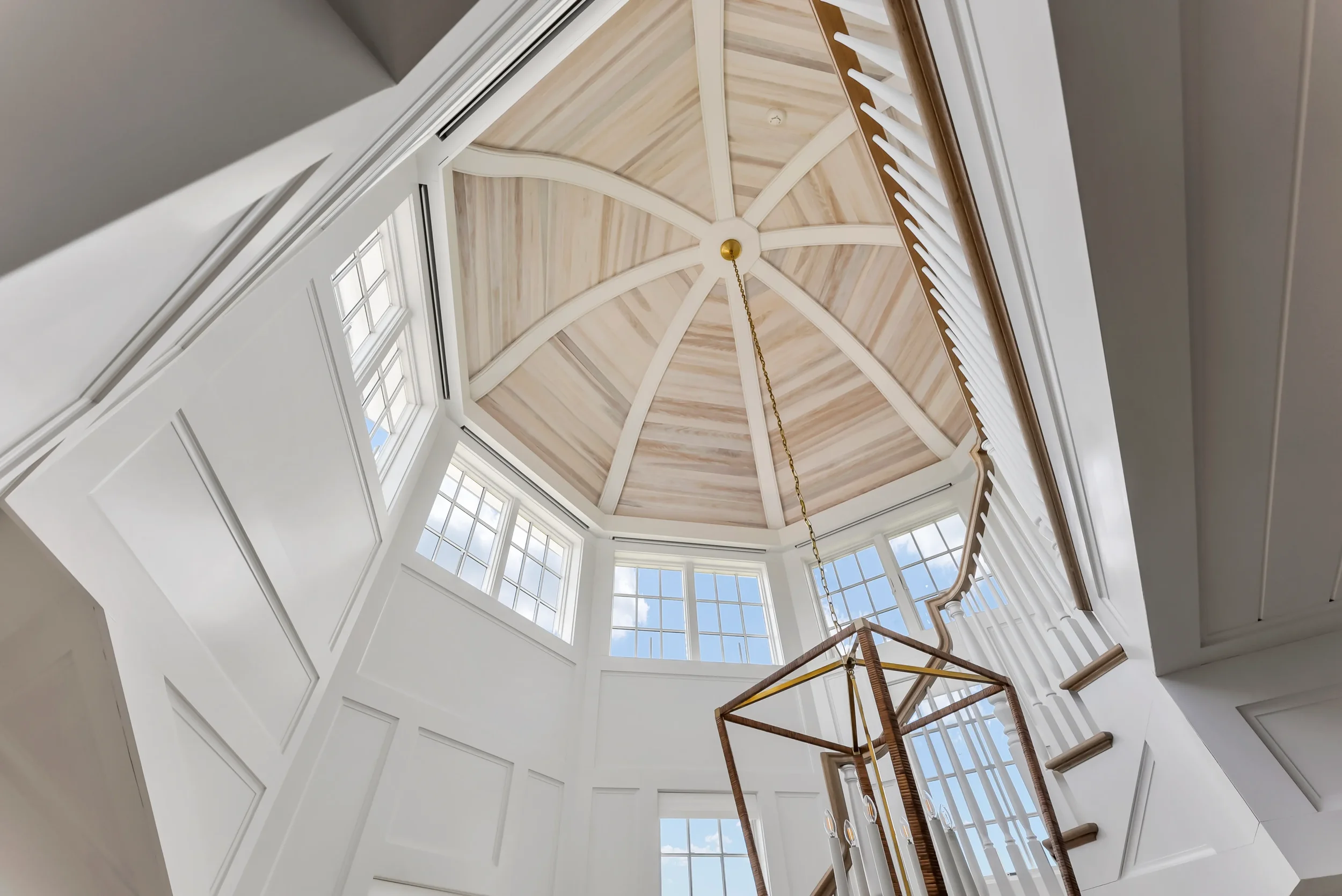 View of a white staircase inside a house looking up towards a wood-paneled octagonal ceiling with large windows and a brass chandelier hanging from the center.