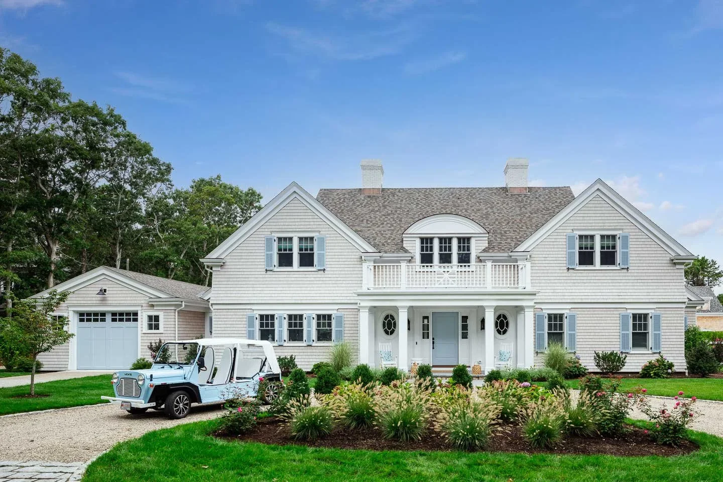 This image showcases a stunning Cape Cod-style estate home that perfectly embodies classic New England architectural elegance. The residence features traditional weathered cedar shingle siding in soft gray tones, complemented by crisp white trim and