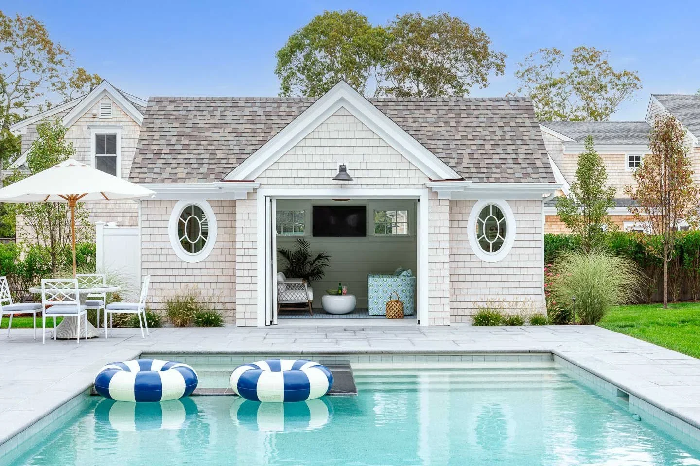 This image showcases a luxurious and inviting outdoor living space that seamlessly blends comfort, style, and natural beauty. The foreground features a covered patio furnished with elegant wicker seating—two armchairs and a sofa—all outfitted with li