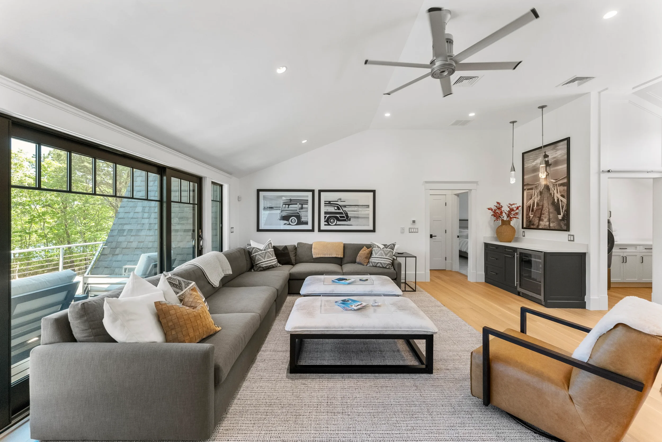 Spacious living room with large sectional sofa, ottomans, armchair, wooden floor, white walls, ceiling fan, large window with balcony, wall art, black cabinet, and sliding glass doors leading outside.