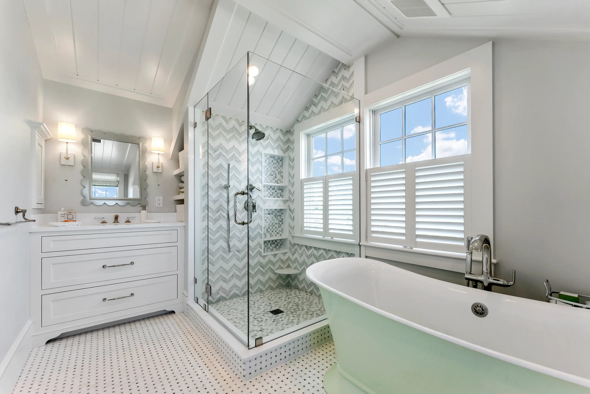 Bright bathroom with a white vanity, a mirror, and sconces, a glass-enclosed shower with patterned tiles, a clawfoot bathtub near windows with shutters, and a white tiled floor.