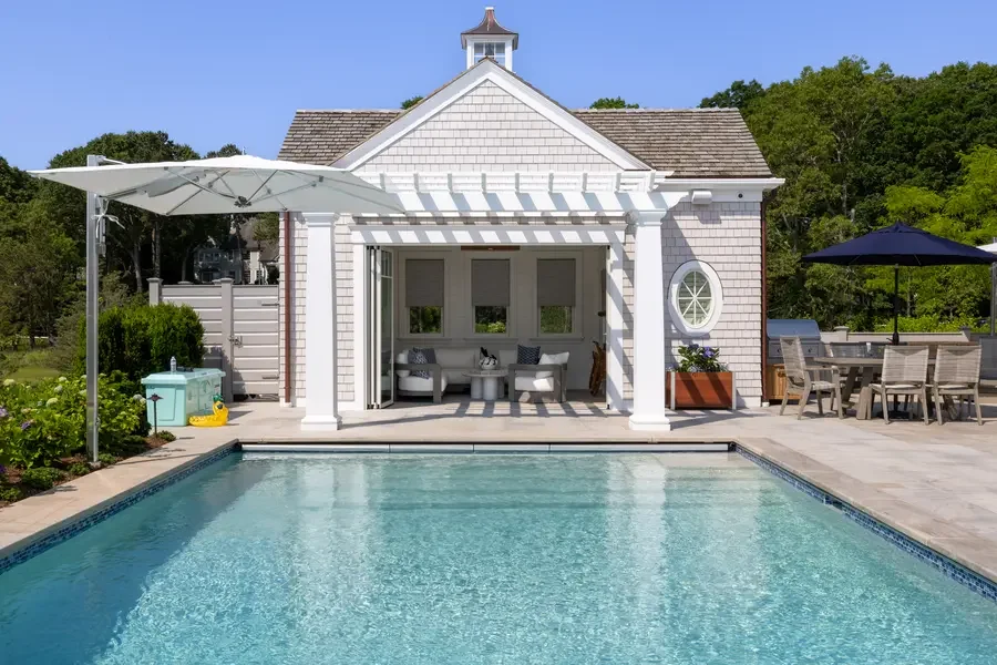 A backyard pool in front of a white pool house with a tiled roof and a cupola. An outdoor dining area with a black umbrella and a seating area with a white sofa inside the pool house.