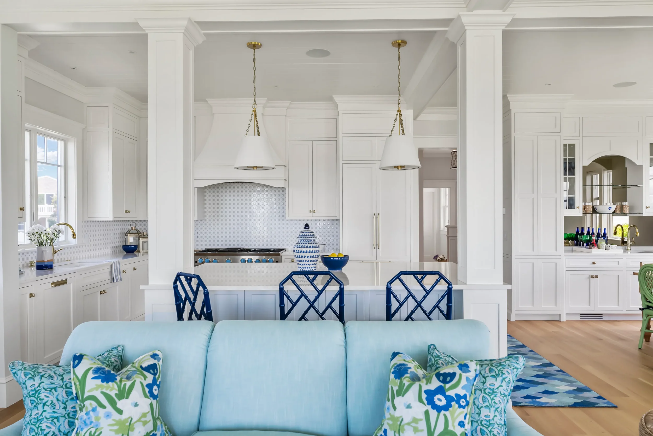 Bright, open kitchen with white cabinetry, gold hardware, and a central island with blue chairs. Decor includes blue bowls, gold faucets, and patterned pillows on a light-colored sofa nearby.