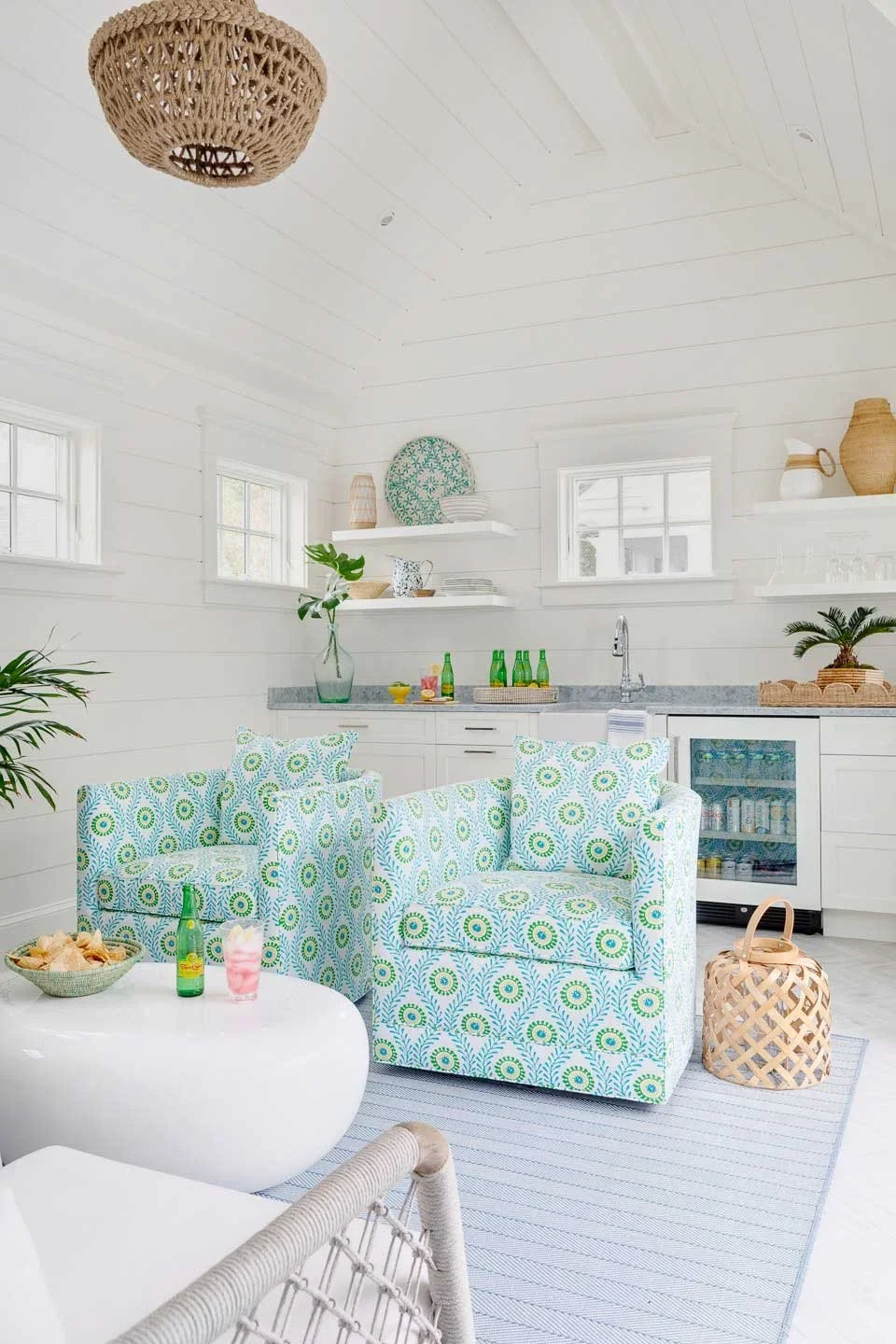 Bright, airy coastal-themed living space with white shiplap walls, patterned armchairs, open kitchen shelves, and tropical decor accents.