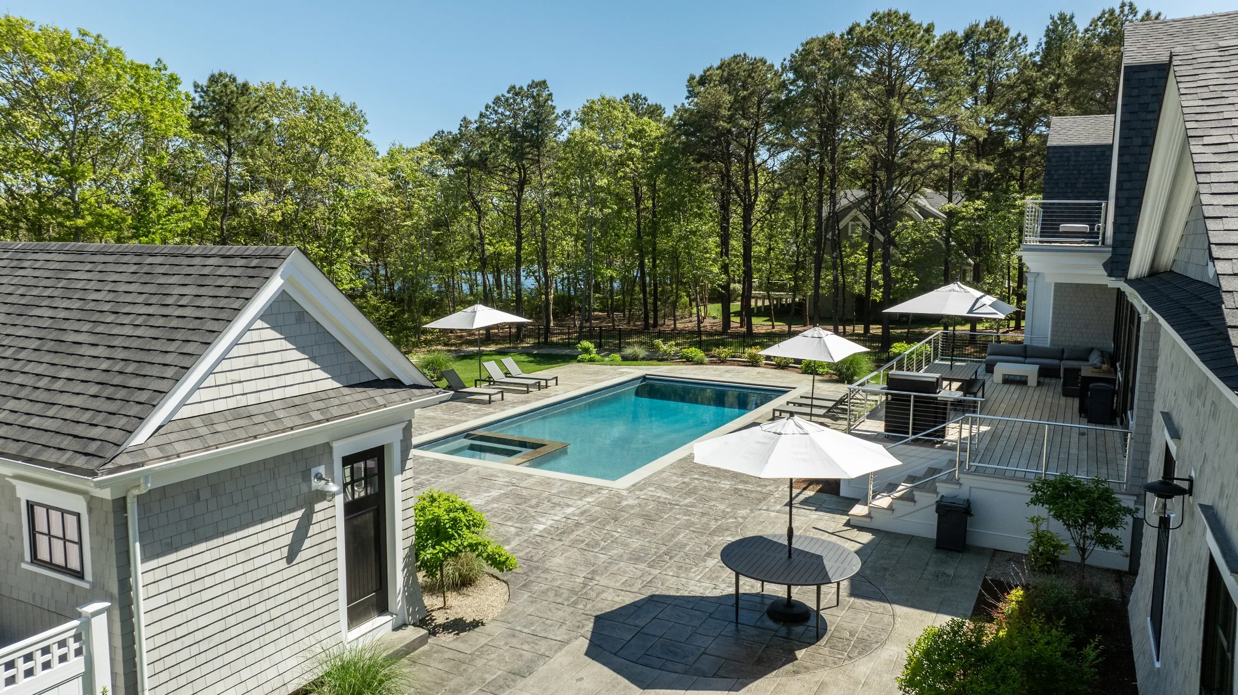 A backyard with a swimming pool, surrounded by patio furniture, umbrellas, and lush green trees.