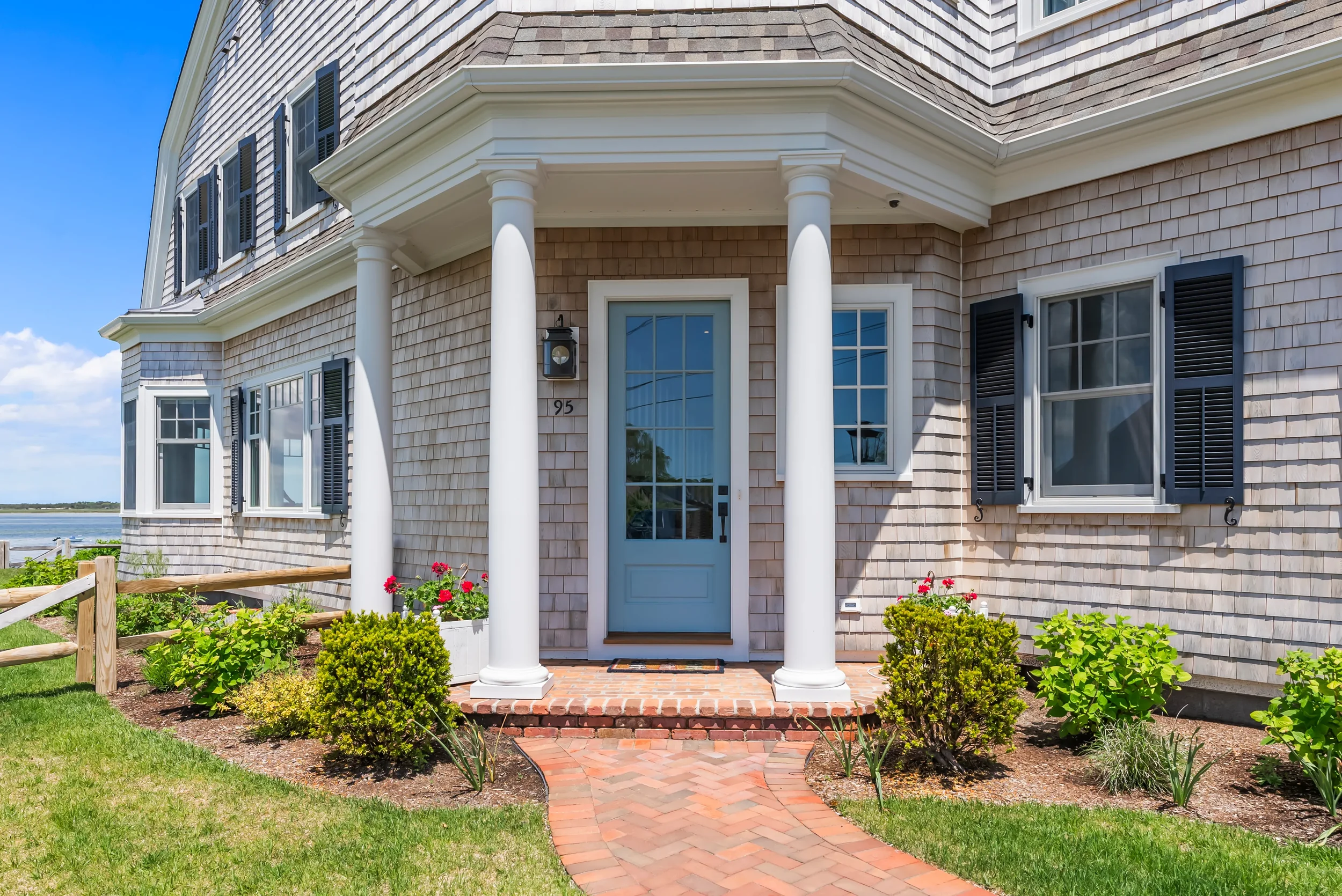 Front view of a house with a blue door, white columns, black shutters, and a brick pathway leading to the porch, surrounded by green grass and plants.