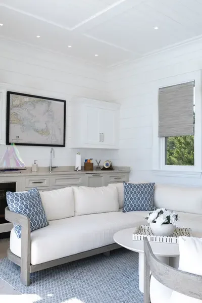 Living room with white sofa, blue patterned pillows, round white coffee table with decorative object, gray upholstered chair, large window with gray Roman shade, and kitchenette in the background with white cabinets and a wall map.
