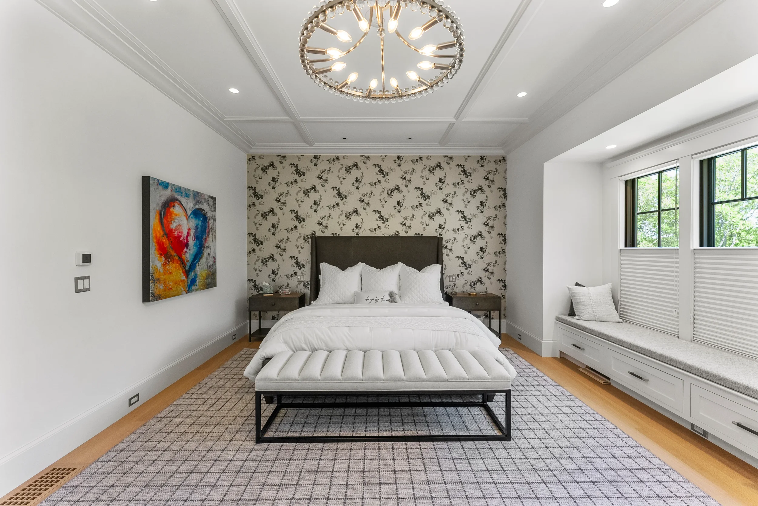 Modern bedroom with white walls, a patterned accent wall behind the bed, and a large circular chandelier. The bed has white bedding and pillows, with a square bench at the foot. There is a window seat with a pillow on the right, and a colorful heart-