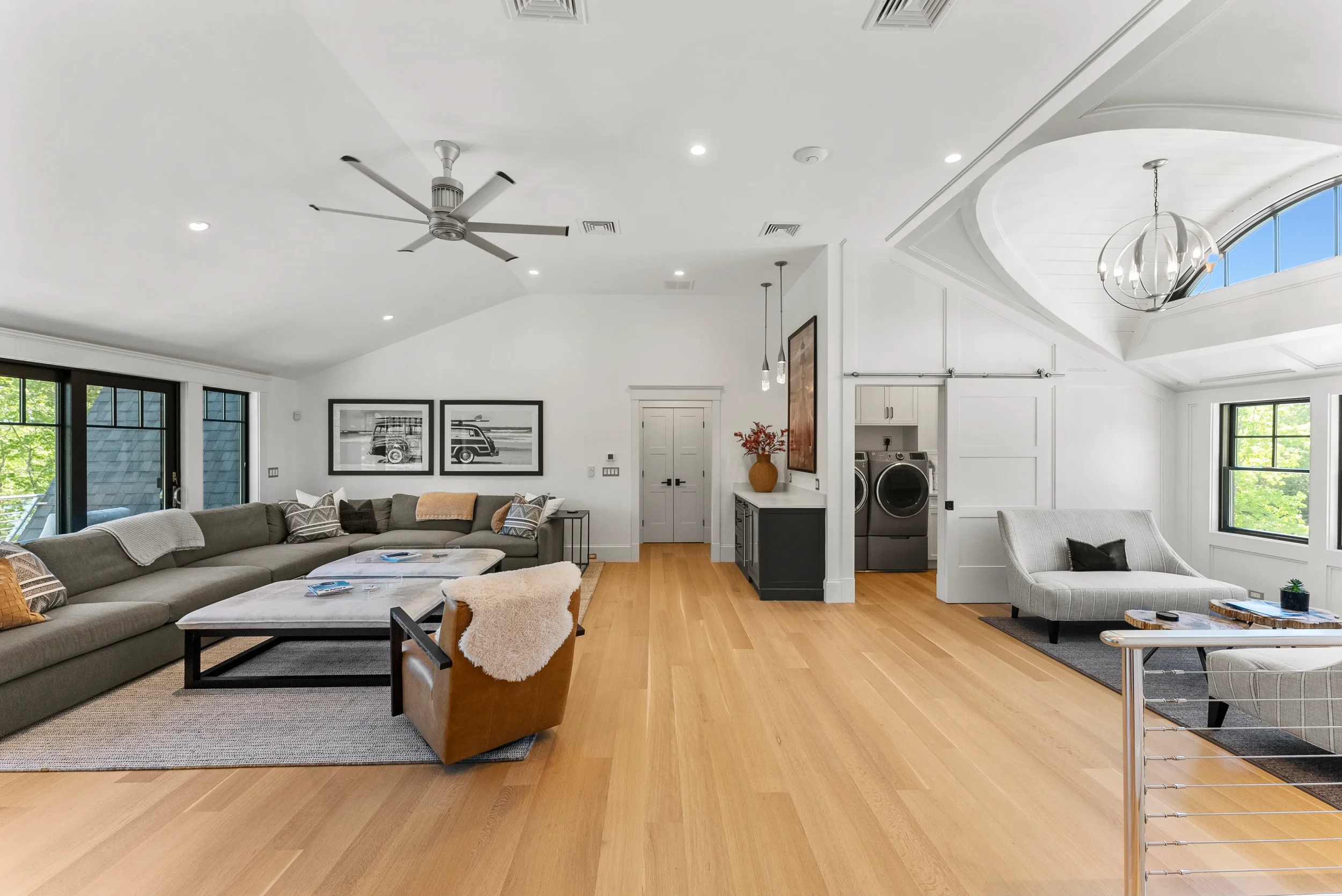 Spacious modern living room with light wood flooring, white walls, multiple seating options including a large sectional sofa and a white armchair, coffee tables, and decorative art. Features large windows and a ceiling fan.