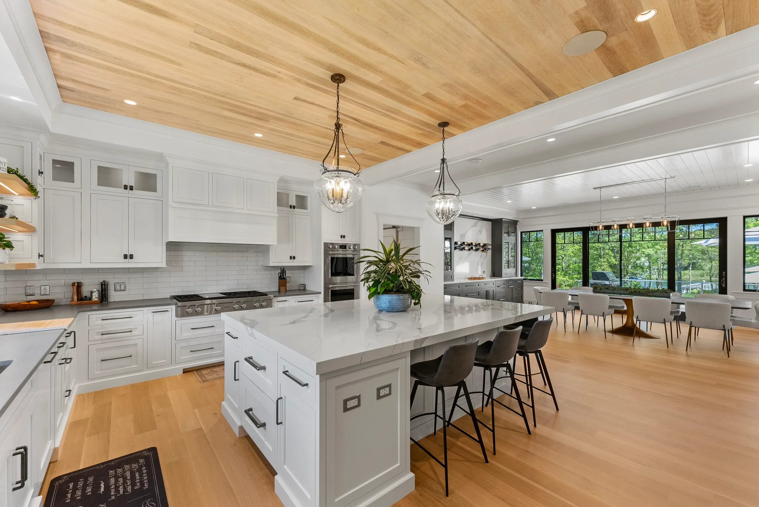 Modern open-concept kitchen and dining area featuring white cabinets, a large island with a marble countertop, wooden ceiling accents, pendant lighting, and large windows overlooking greenery.