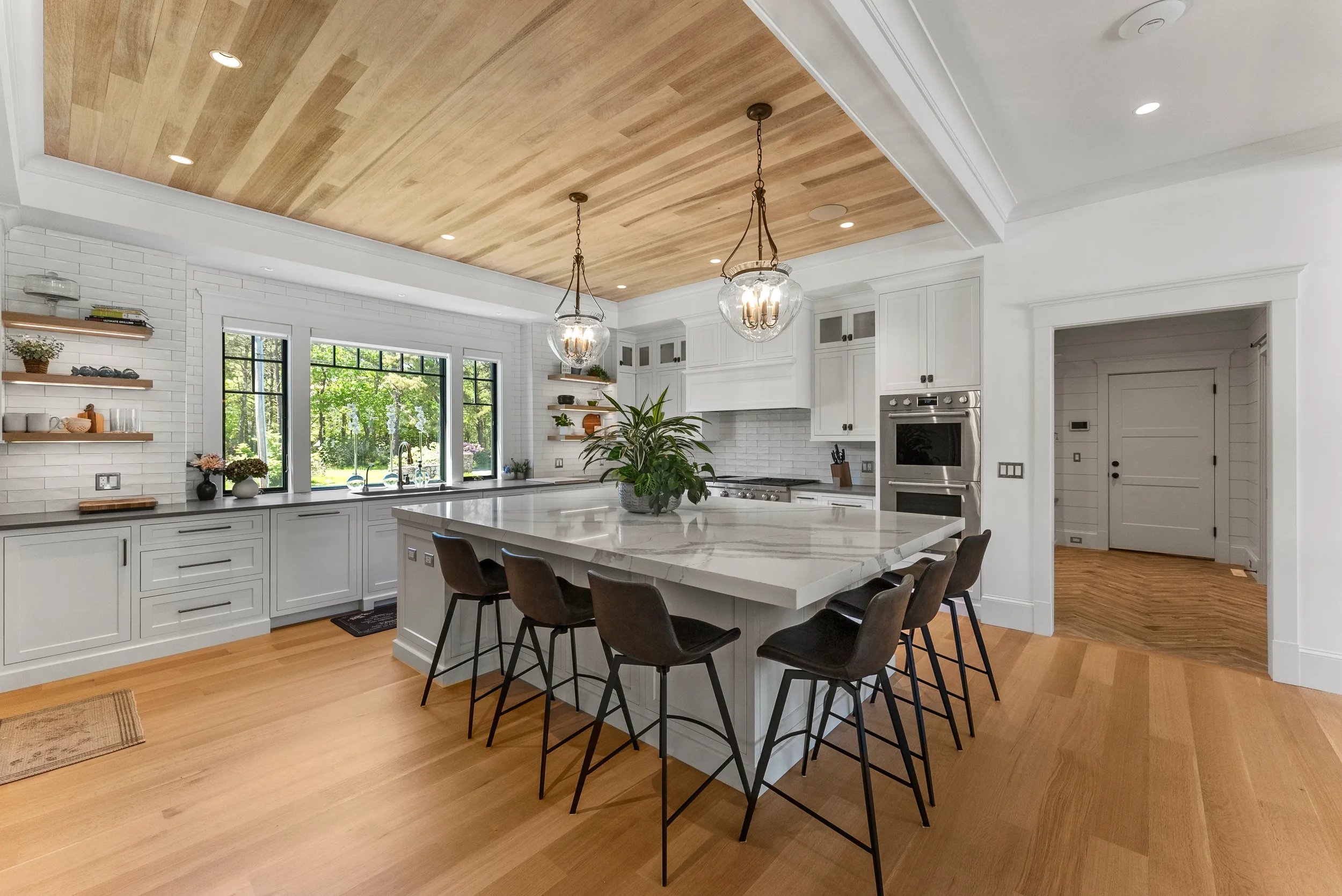 Modern kitchen with white cabinets, a large marble island, black chairs, wooden ceiling, and large window with green view.