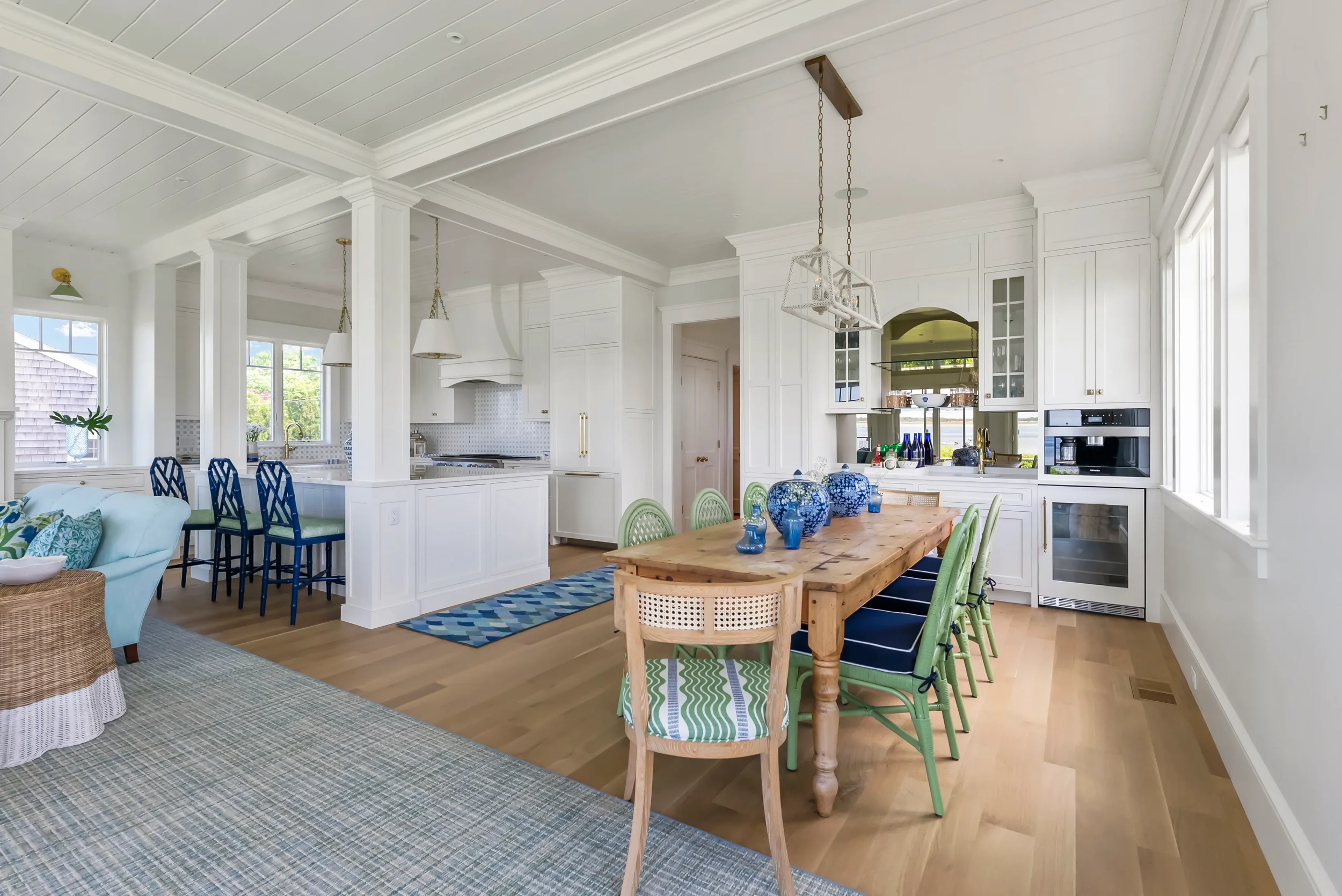 Bright open-concept kitchen and dining area with white cabinetry, hardwood floors, and large windows. Features a wooden dining table with green and navy chairs, a white sectional sofa, and blue decorative vases.