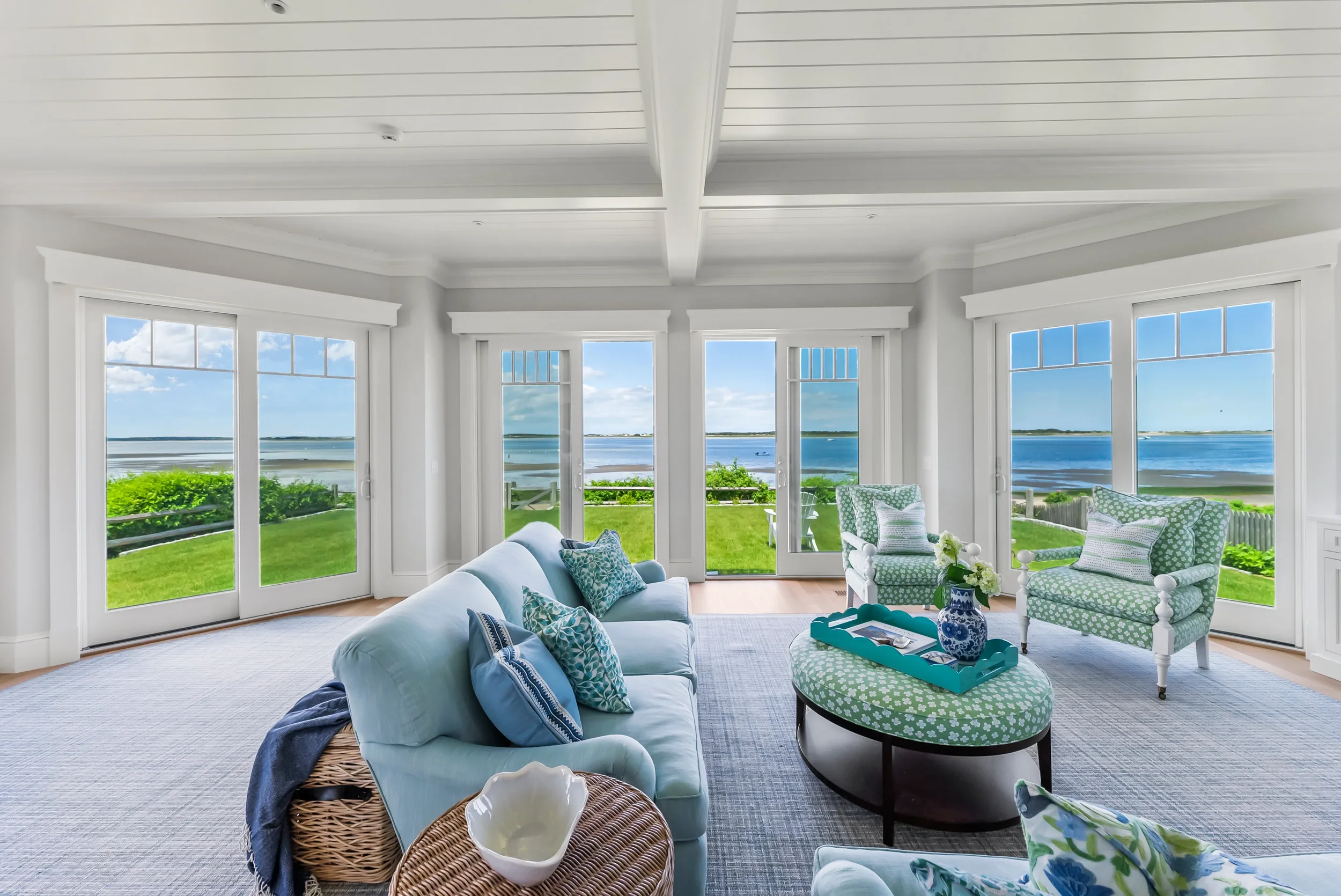 Living room with large windows showcasing water view, white walls, white ceiling with beams, light blue sofa with patterned pillows, two patterned armchairs, oval ottoman with tray and vase, and a small wicker side table.