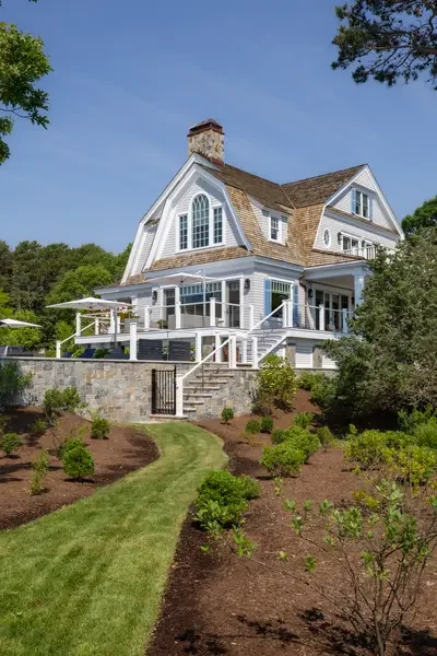 A large white house with a steep roof, multiple gables, and large windows, surrounded by green trees and a landscaped yard with a stone retaining wall and a staircase leading to the house.