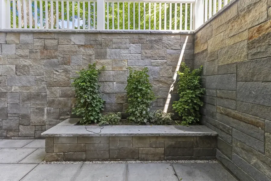 This image showcases a sophisticated outdoor living space featuring a modern built-in stone bench that serves as both seating and a planter. The bench is constructed from the same natural stone blocks used throughout the space, creating a cohesive de
