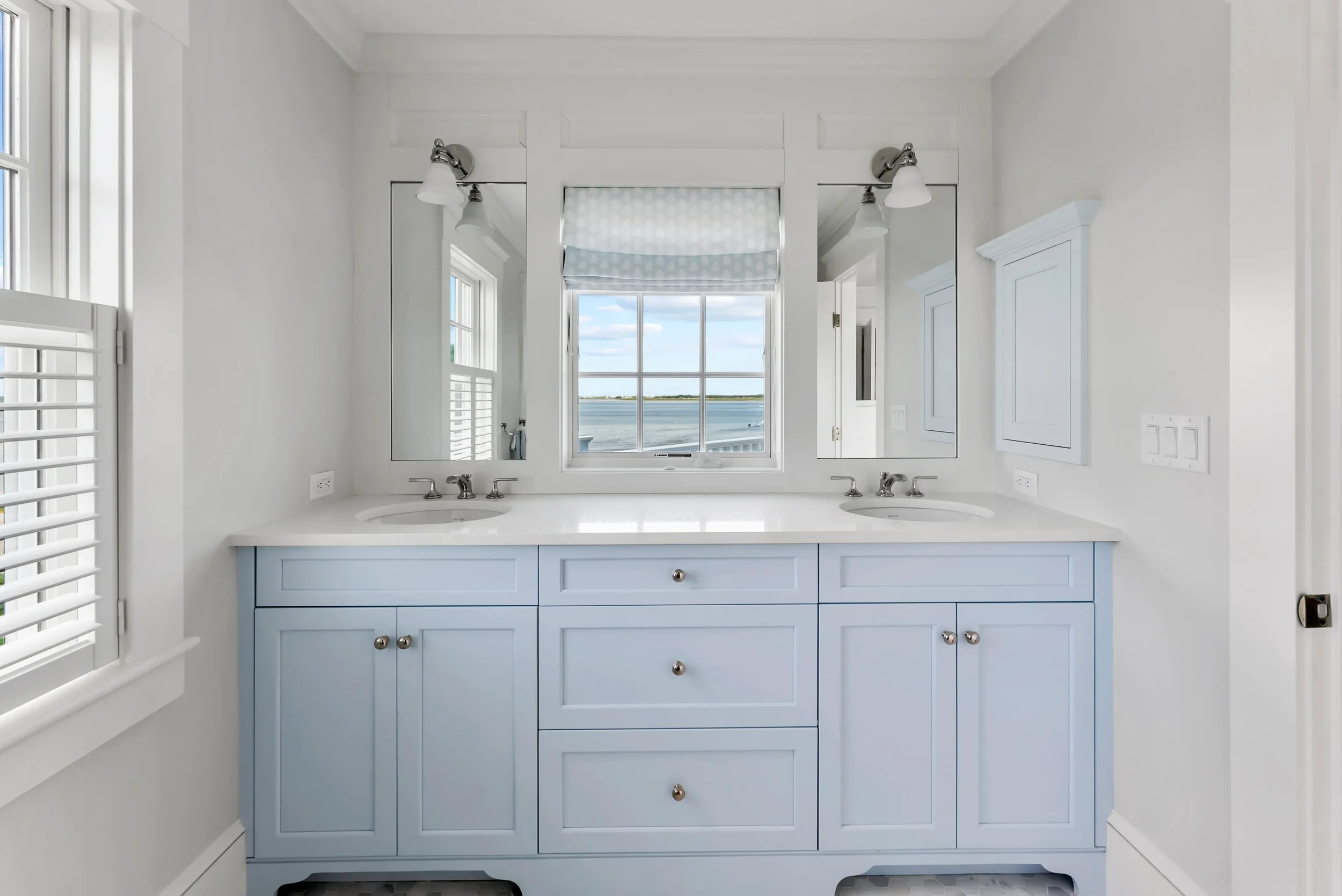 A bright bathroom with a double vanity featuring light blue cabinets, two sinks, and two mirrors. A window over the vanity offers a view of a body of water and the sky.