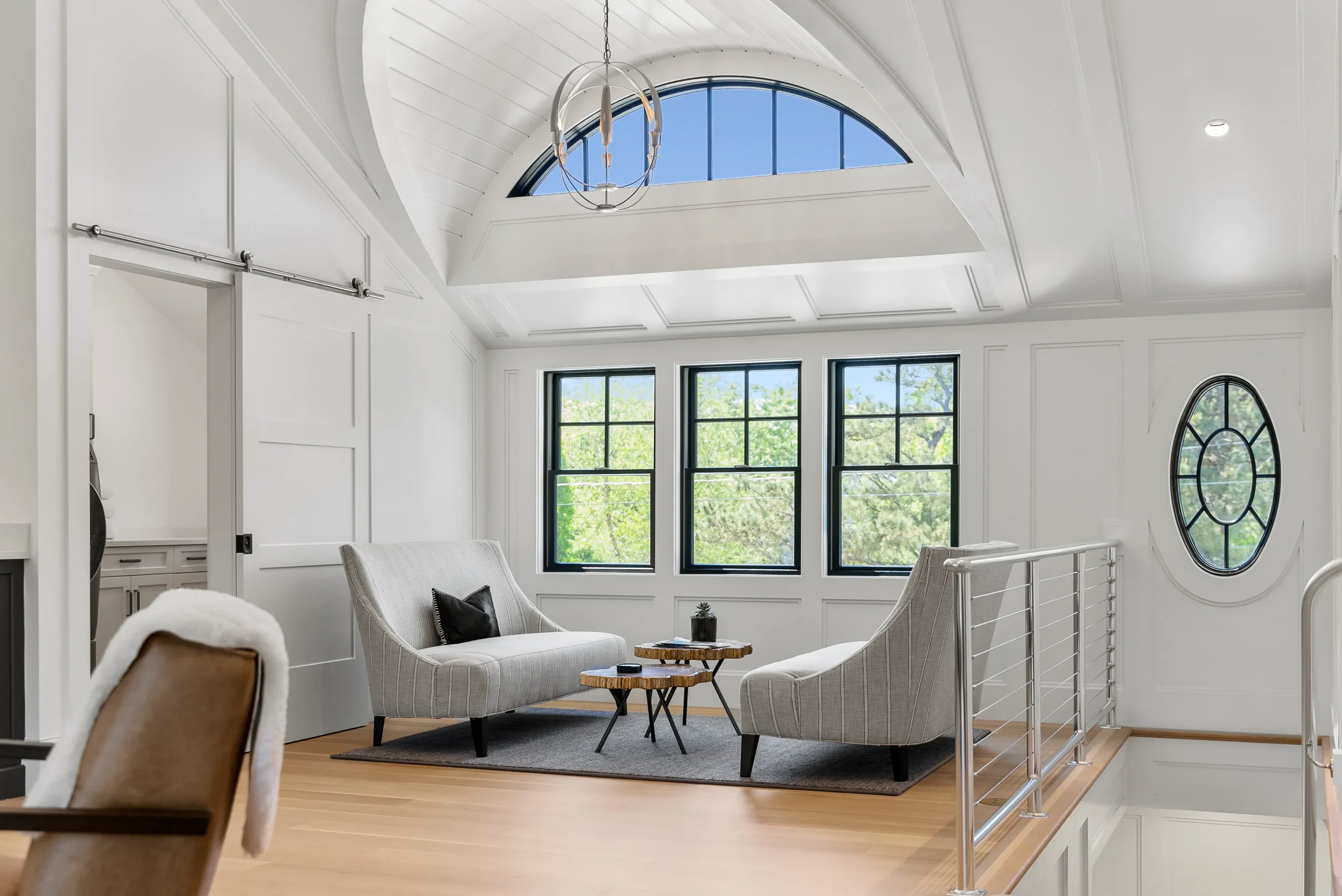 Bright, modern living room with white walls, large windows, a vaulted ceiling, a hanging chandelier, and two light-colored armchairs arranged around a small coffee table.