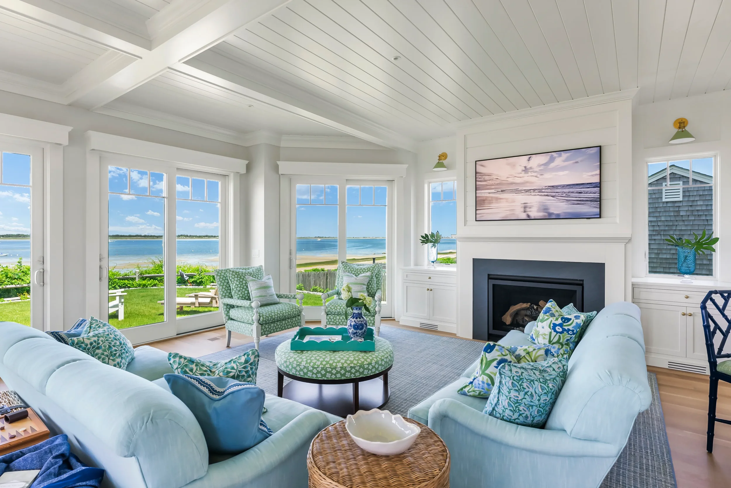 Bright living room with large windows overlooking a lake, white walls, a white fireplace, a mounted TV, and green and blue upholstered furniture.