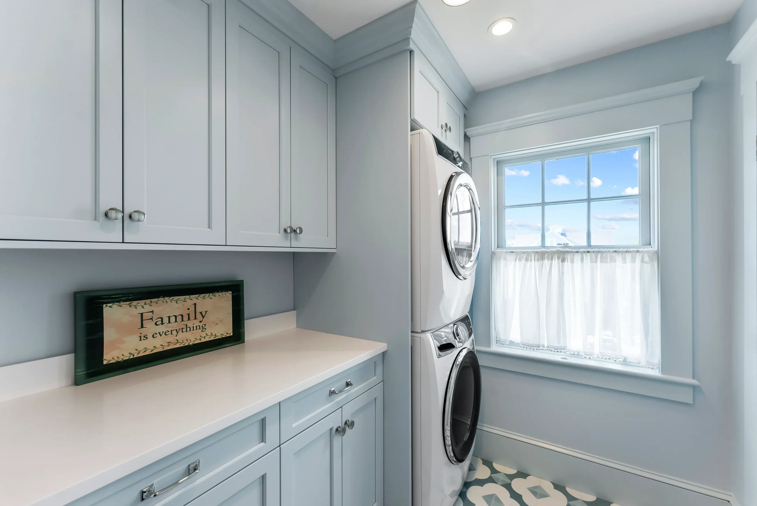 Laundry room with stacked washer and dryer, pale blue cabinets, a window with white curtains, and a sign that says 'Family is everything' on the counter.