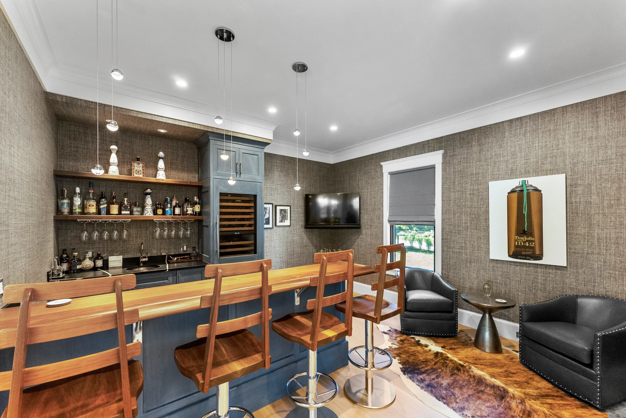 Living room with bar area, two black armchairs, a small side table, a large window with a gray shade, a wall-mounted flat-screen TV, and wall art depicting a brown liquor bottle. The room has beige textured walls, crown molding, and a cowhide rug.
