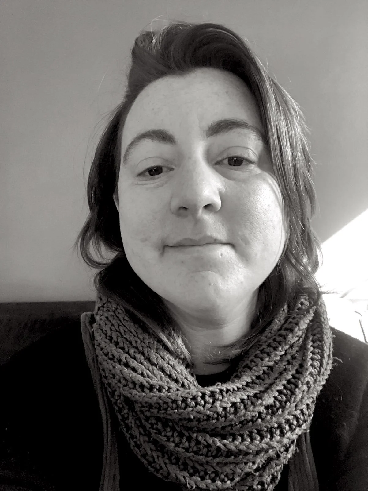A close up black and white photo of Soph. They are warmly looking at the camera, wearing a cozy knit scarf