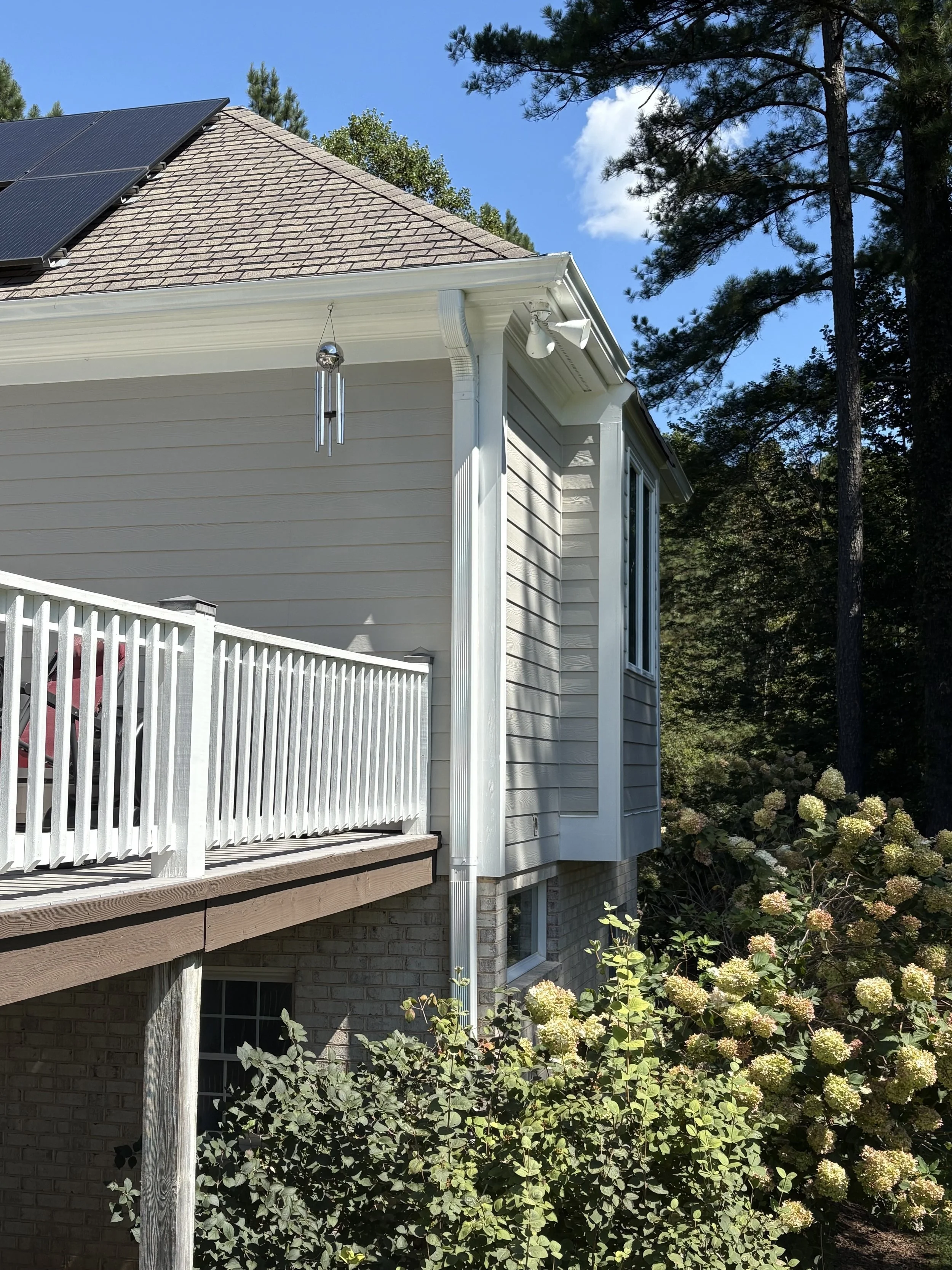 Side view of a house showing a roof with solar panels, beige siding, white trim, a balcony with a white railing, and lush bushes with white flowers, under a clear blue sky.
