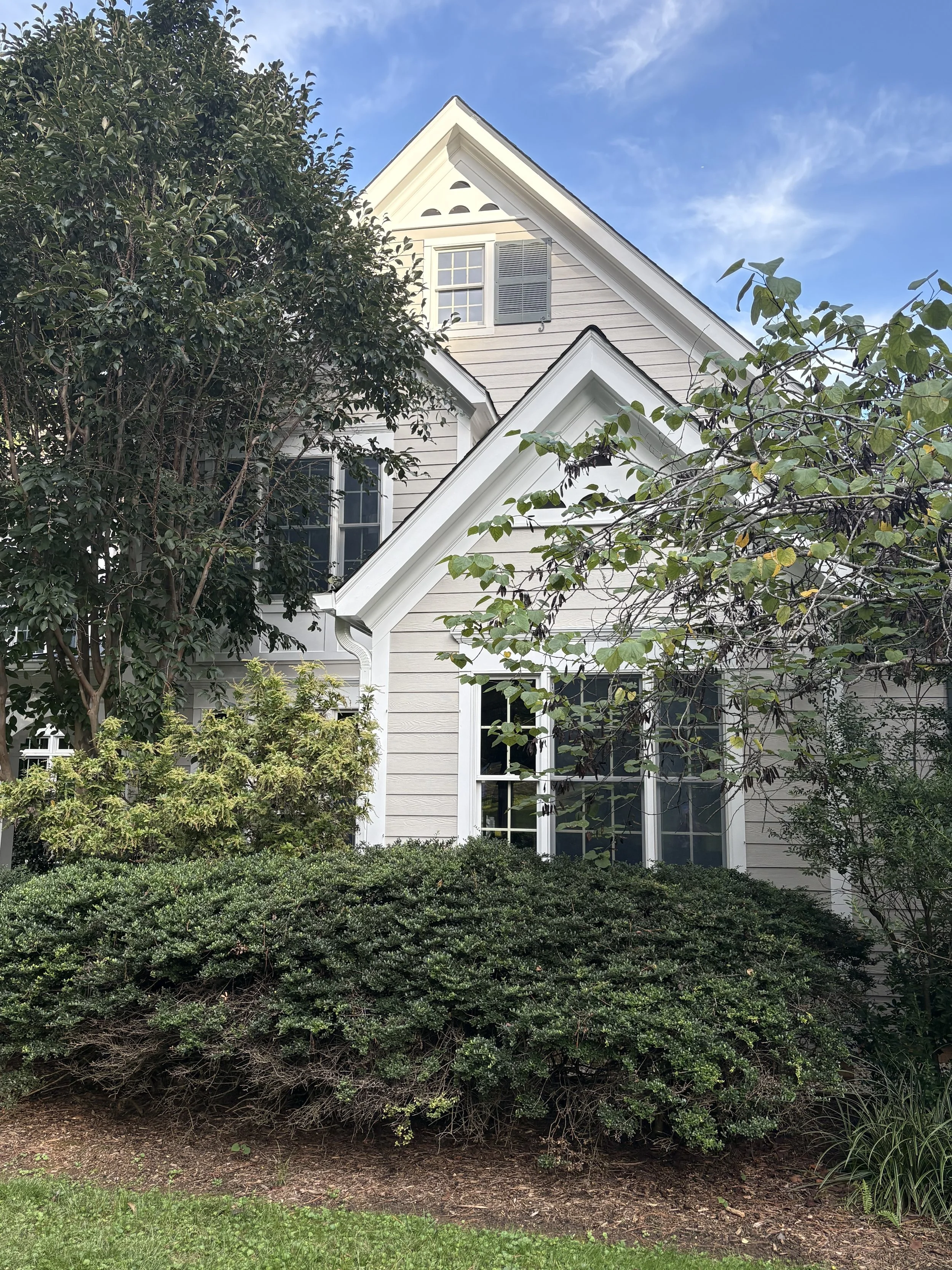 A house with gray siding, white trim, and multiple windows, surrounded by green bushes and trees under a partly cloudy blue sky.