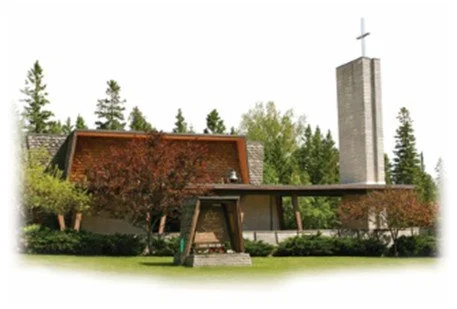Modern church building with a tall cross on a spire, surrounded by trees and greenery.
