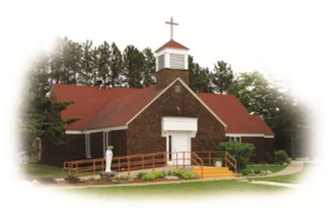 A brick church with a cross on top, red roof, and white entrance door, surrounded by trees and a walkway with a person standing outside.