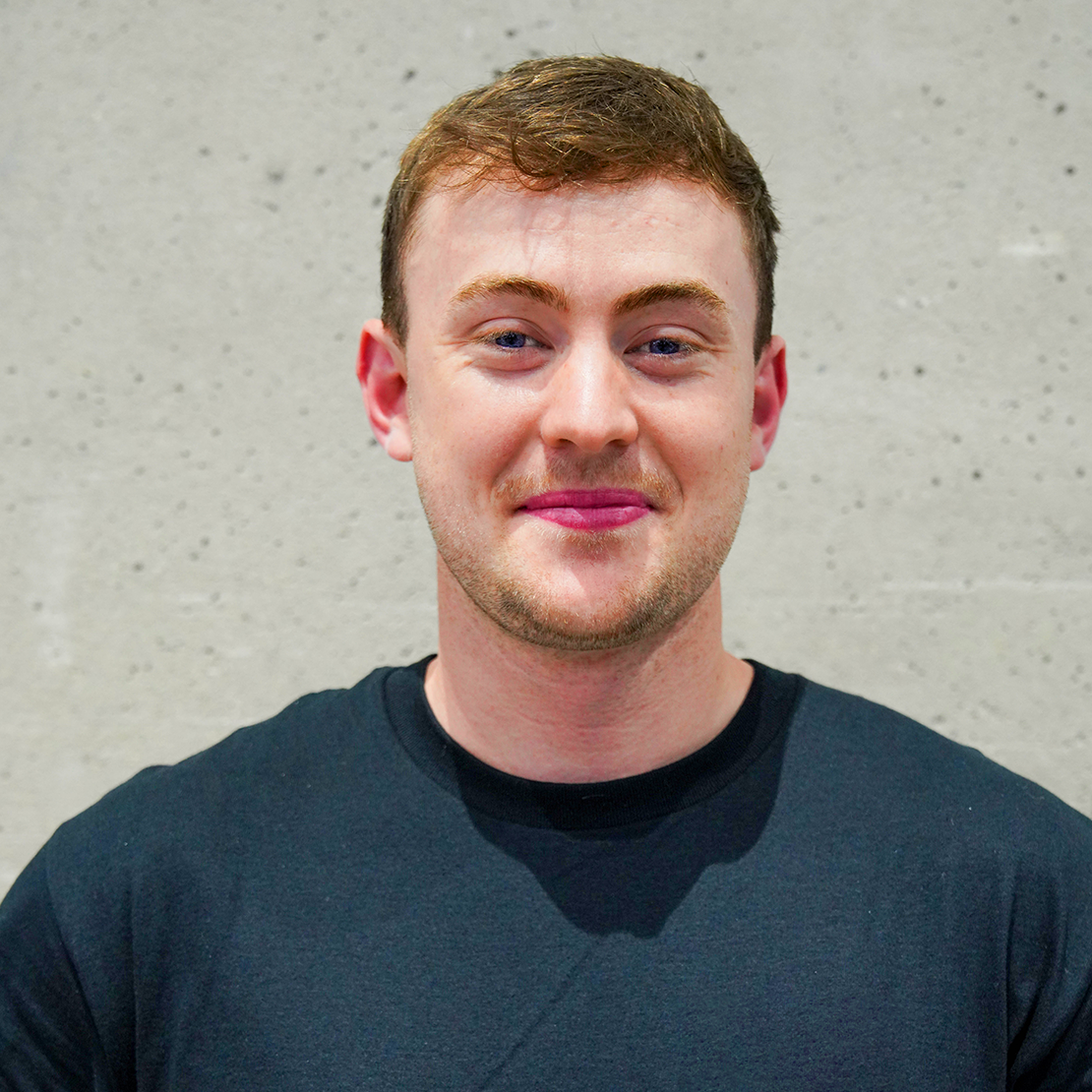 A young man with short brown hair, fair skin, wearing a black t-shirt, standing in front of a gray textured wall.