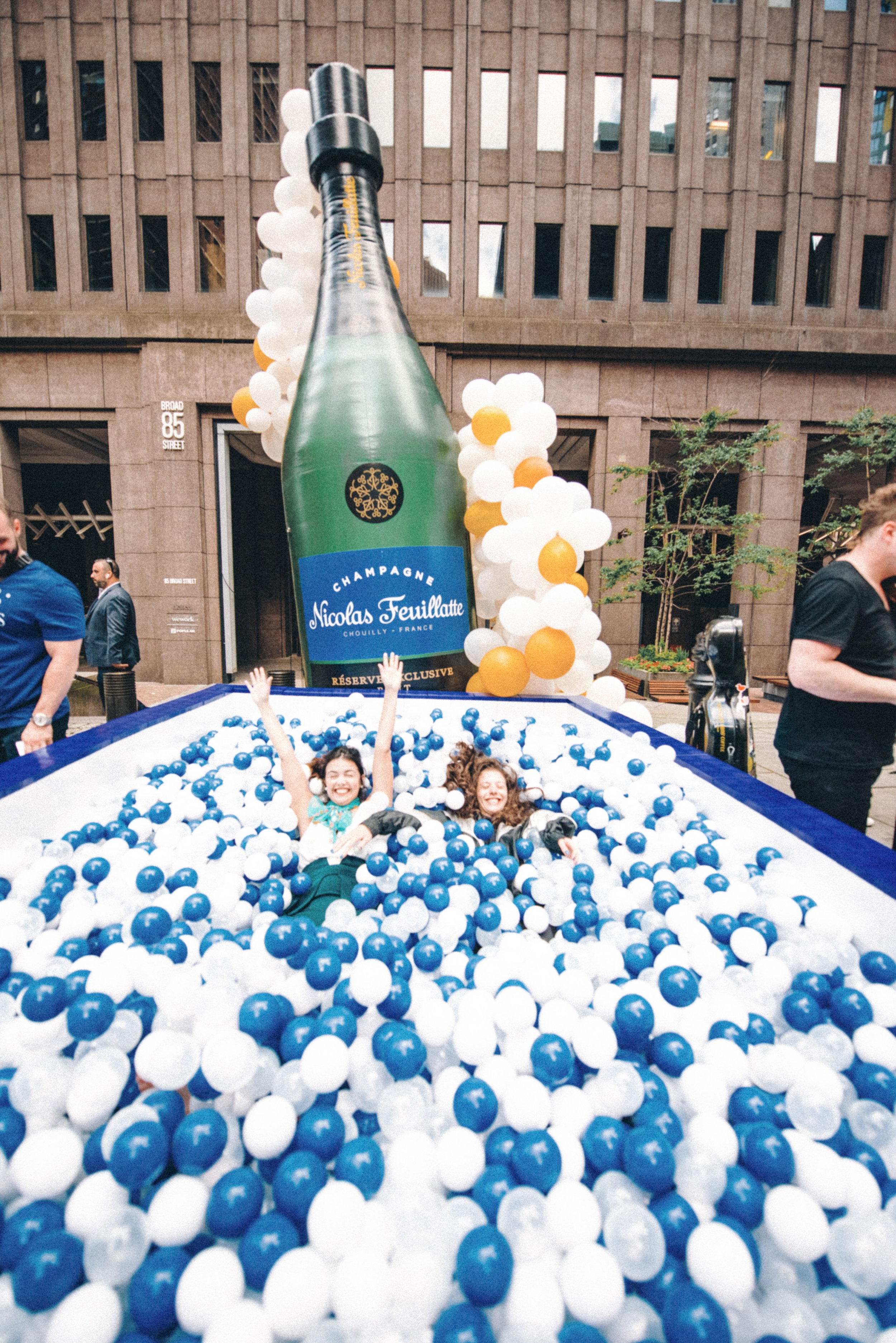 Two women smiling and splashing in a large ball pit filled with blue and white plastic balls, with a giant inflatable champagne bottle decoration behind them.