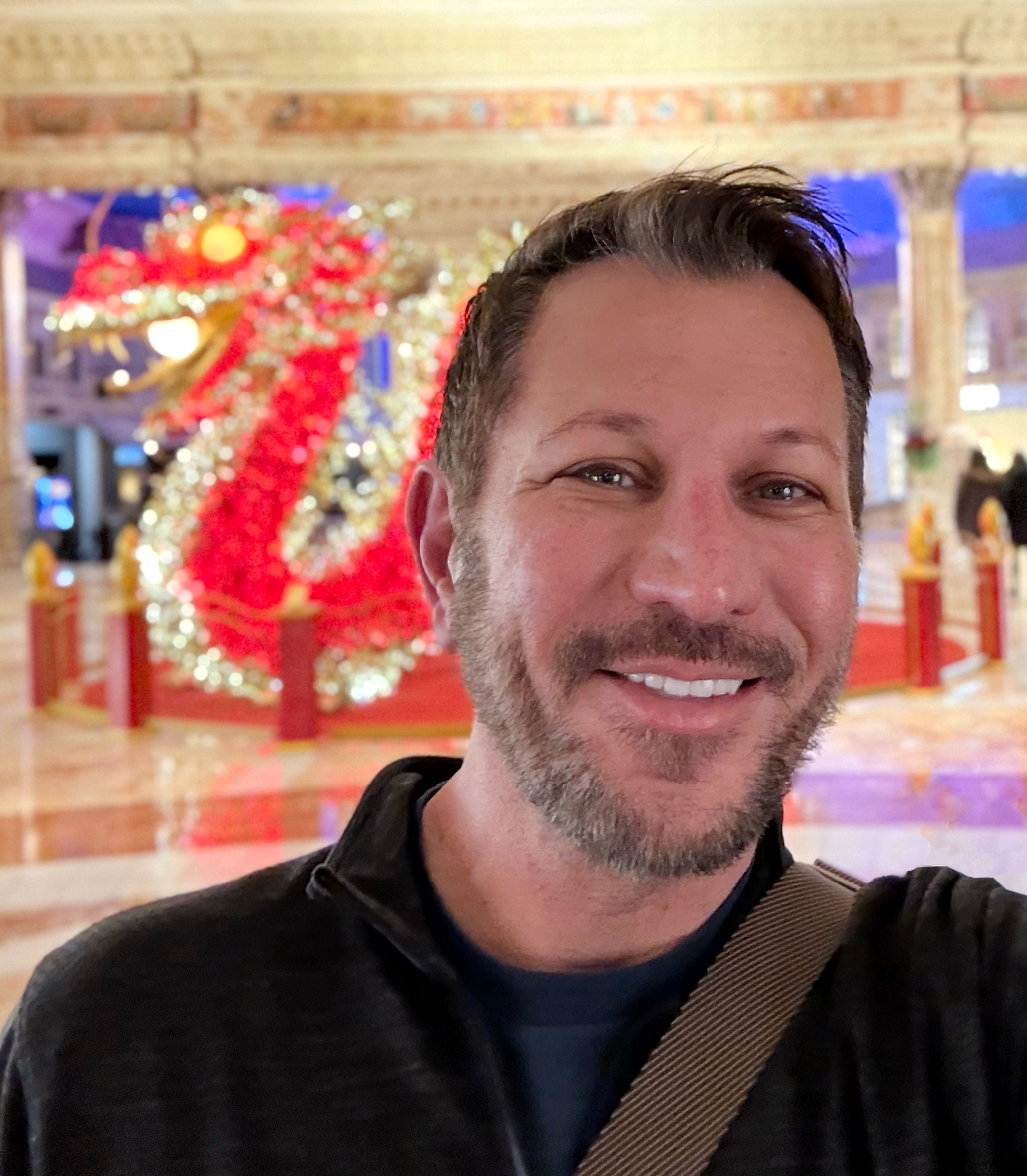 A man with short brown hair and a beard, wearing a black long-sleeve shirt, standing confidently with arms crossed on a busy city street during daytime.