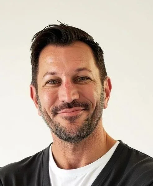 A man with short brown hair and a beard, wearing a black long-sleeve shirt, standing confidently with arms crossed on a busy city street during daytime.