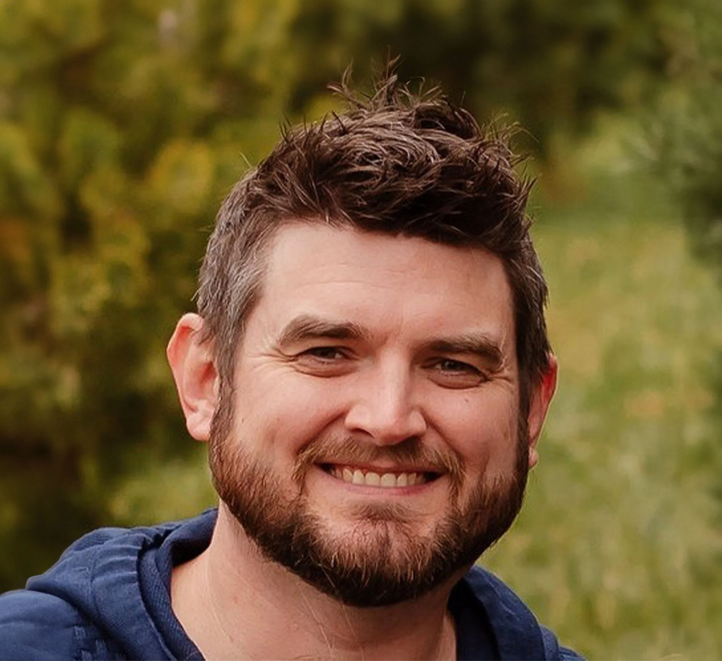 A smiling man with brown hair and beard outdoors with blurred green trees in the background.